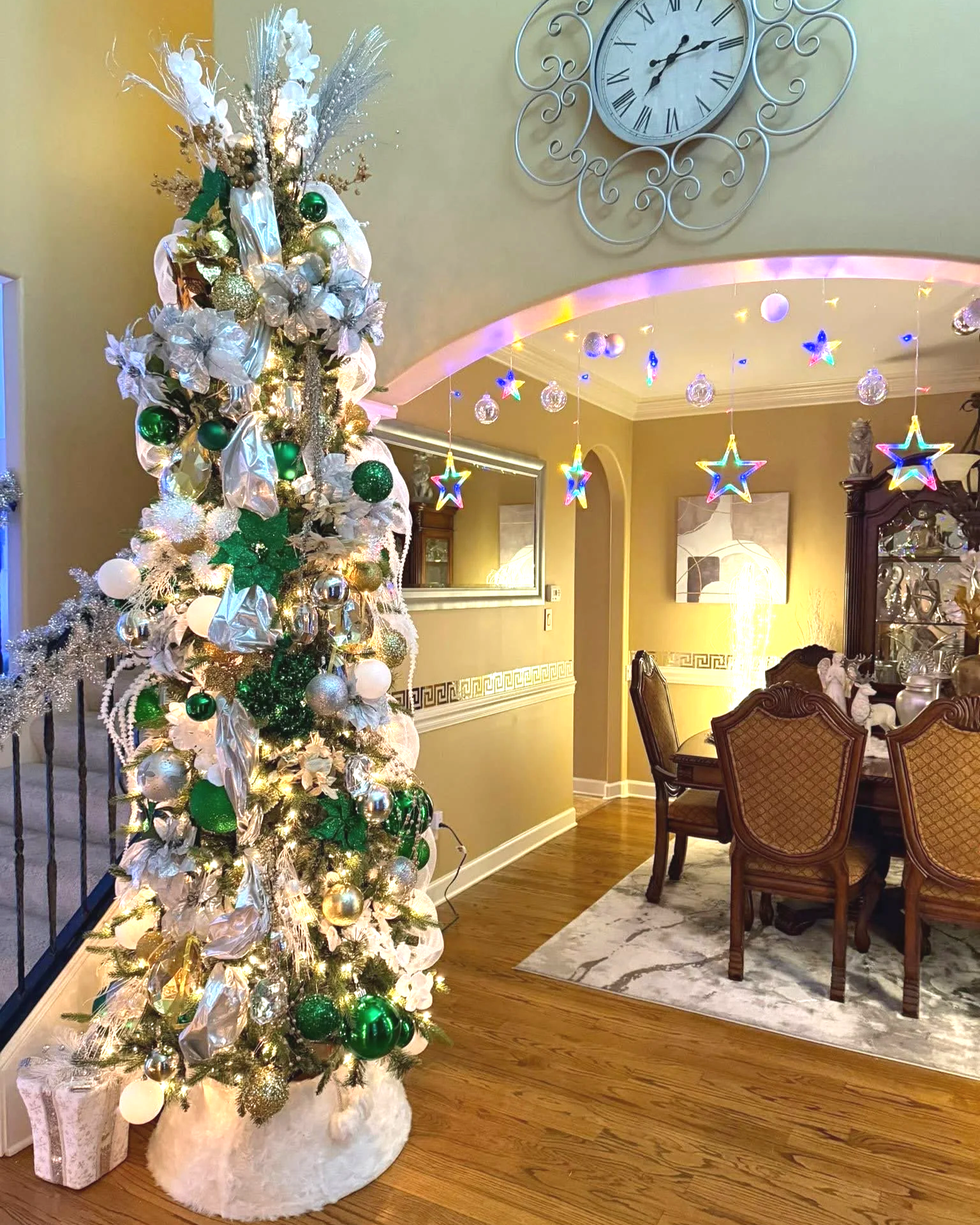Decorated Christmas tree with gold, white, and green ornaments, ribbon accents, and lights in a living room. In the background, there are star-shaped and ball-shaped hanging lights, a dining table with chairs, a marble rug, a wall mirror, and a clock on the wall.