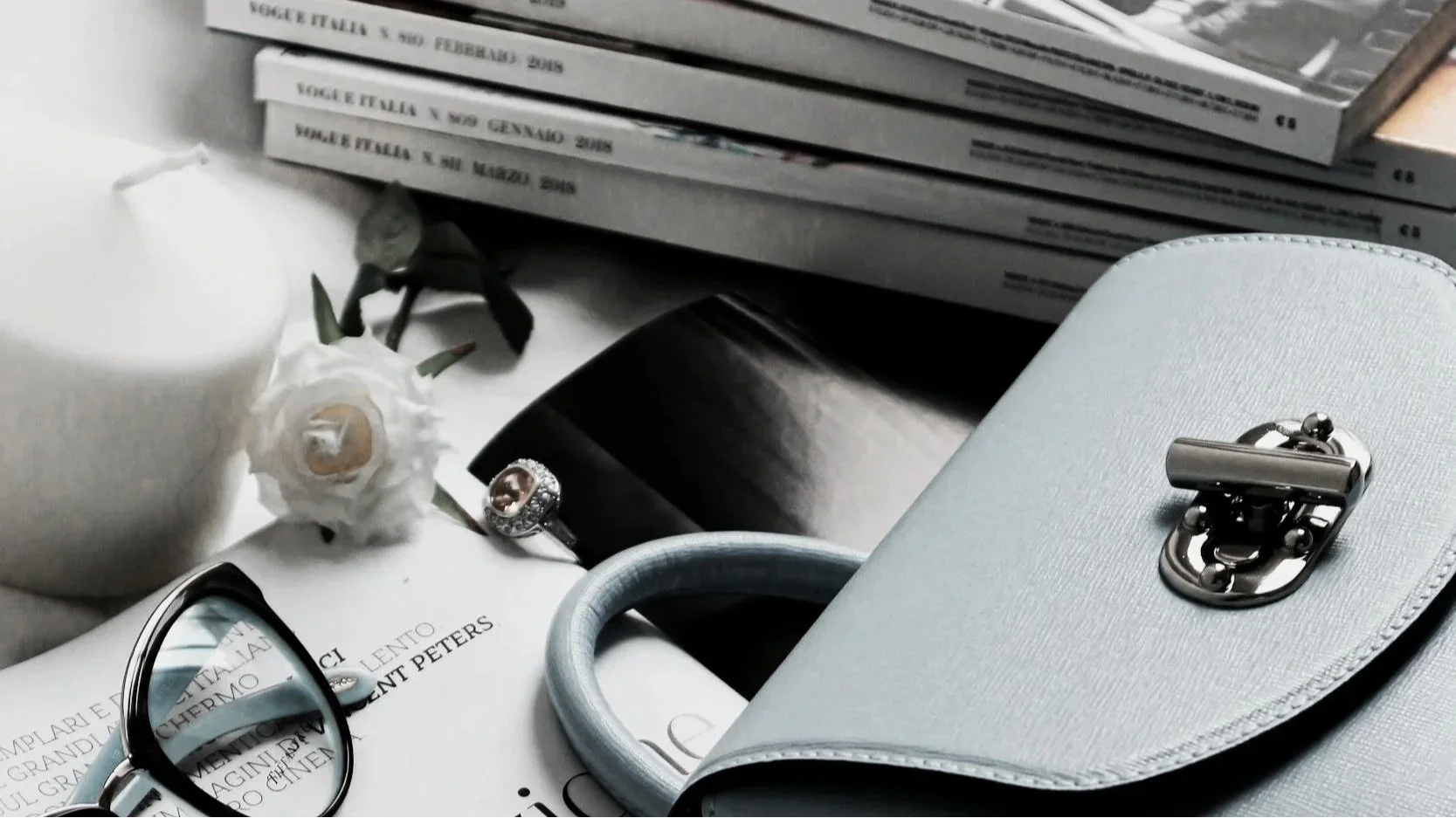 A flat lay of various objects including a gray handbag, a pair of eyeglasses, a white flower, a ring, a rolled white towel, a candle, and magazines or books with Italian titles.