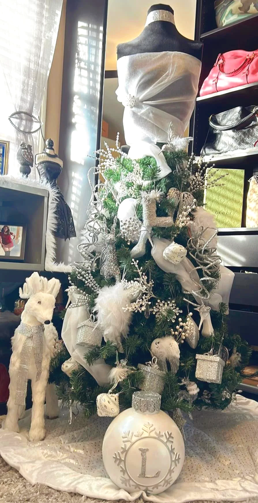 Decorated Christmas tree with white ornaments, including animals, presents, and a large ornament with the letter 'L'. A white plush llama figurine stands nearby. A mannequin draped with white fabric is behind the tree, with a shelf of handbags in the background.