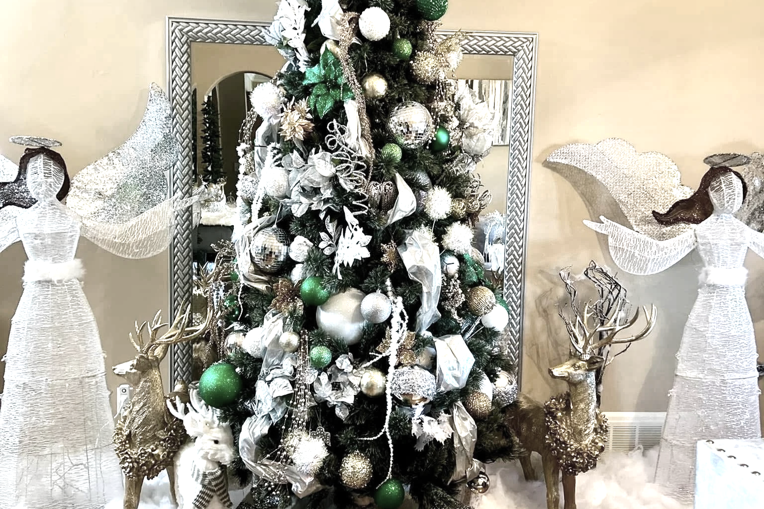 A Christmas tree decorated with white, gold, and green ornaments, surrounded by two angel figurines with large wings and gold reindeer figurines.