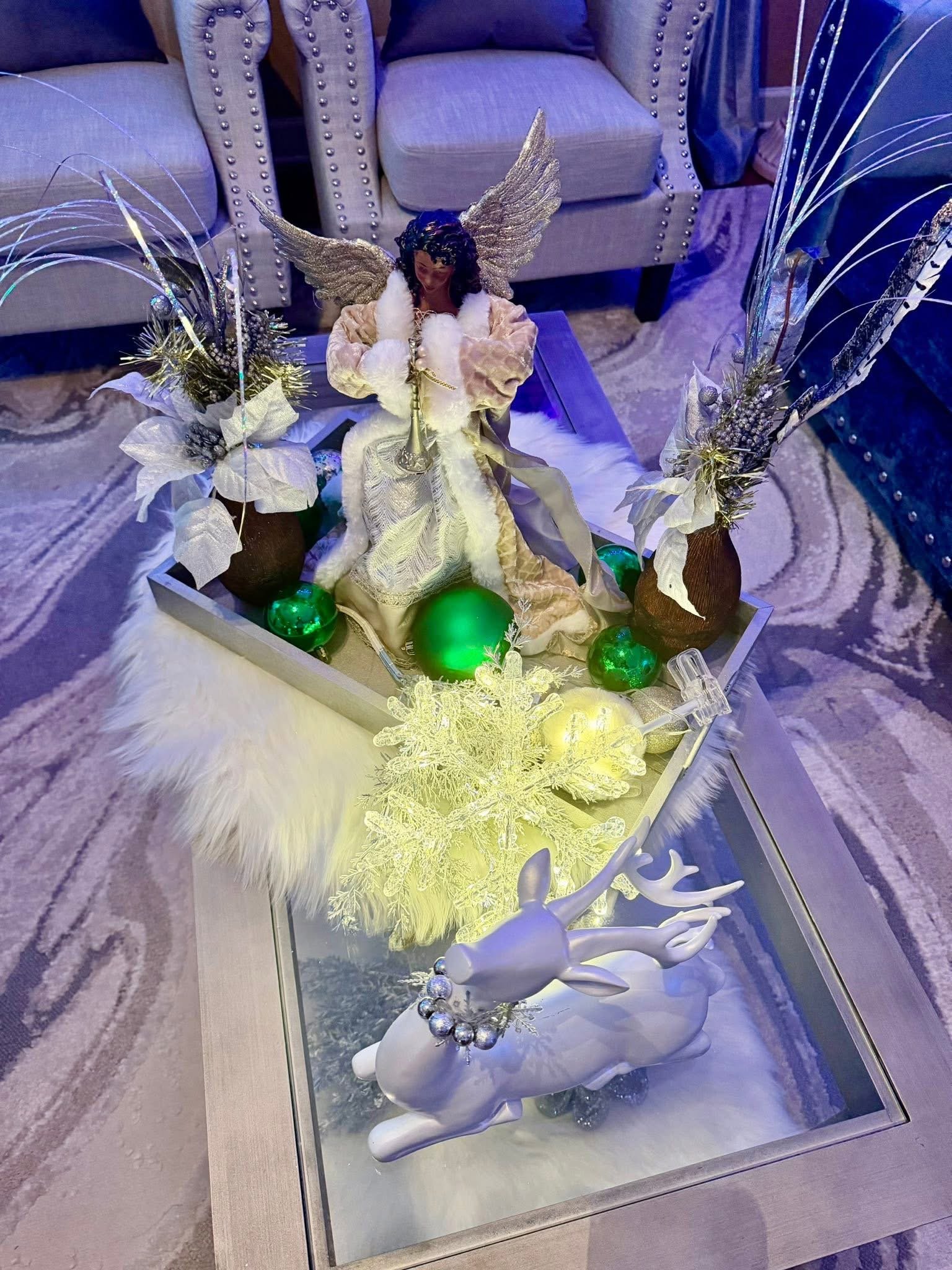 Decorative holiday display featuring an angel figurine with wings, surrounded by green ornaments, white poinsettia flowers, and illuminated decorative snowflakes, with a white reindeer figurine in the foreground on a glass table.
