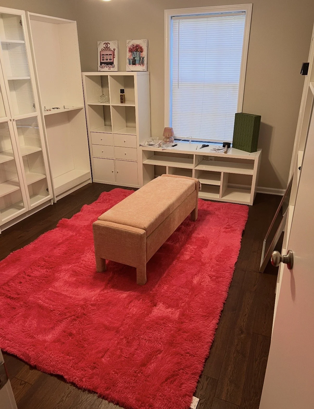 Empty room with a red shaggy rug, a pink upholstered bench, white shelving units, a window with closed blinds, and various small items on the shelves and window sill.