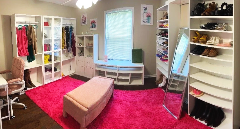 A walk-in closet with white shelving units filled with clothing, shoes, and accessories. A pink rug covers part of the dark wood floor, and a pale pink ottoman sits in the center. There is a window with a light blue blind, and a full-length mirror leaning against the shelves.