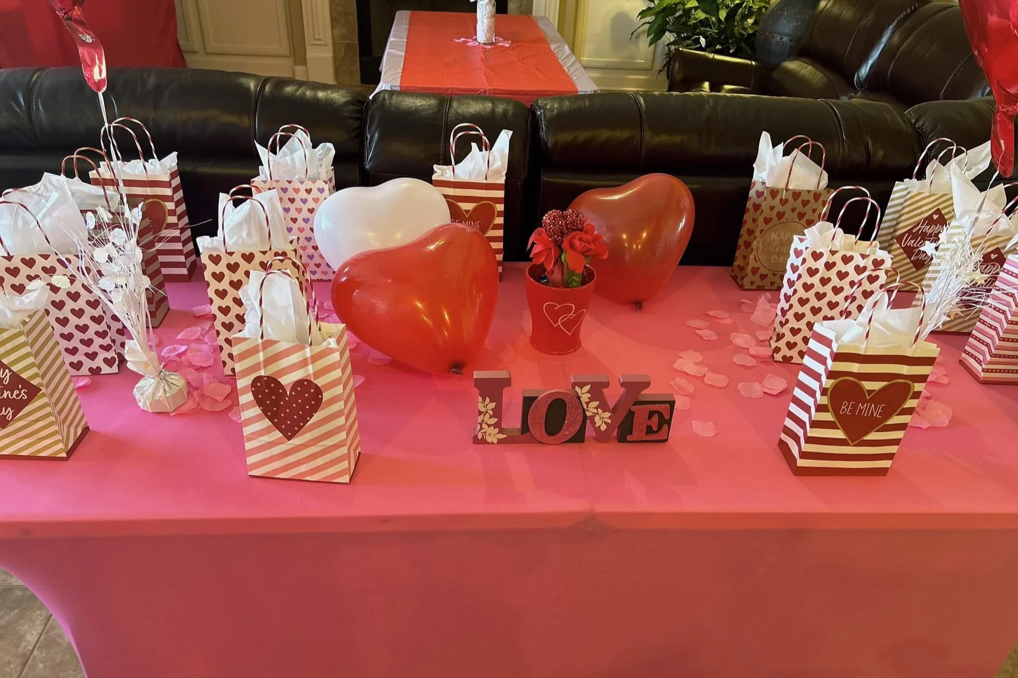 Valentine's Day themed table decorated with pink tablecloth, heart-shaped balloons, gift bags with hearts and stripes, and the word LOVE spelled out on the table.