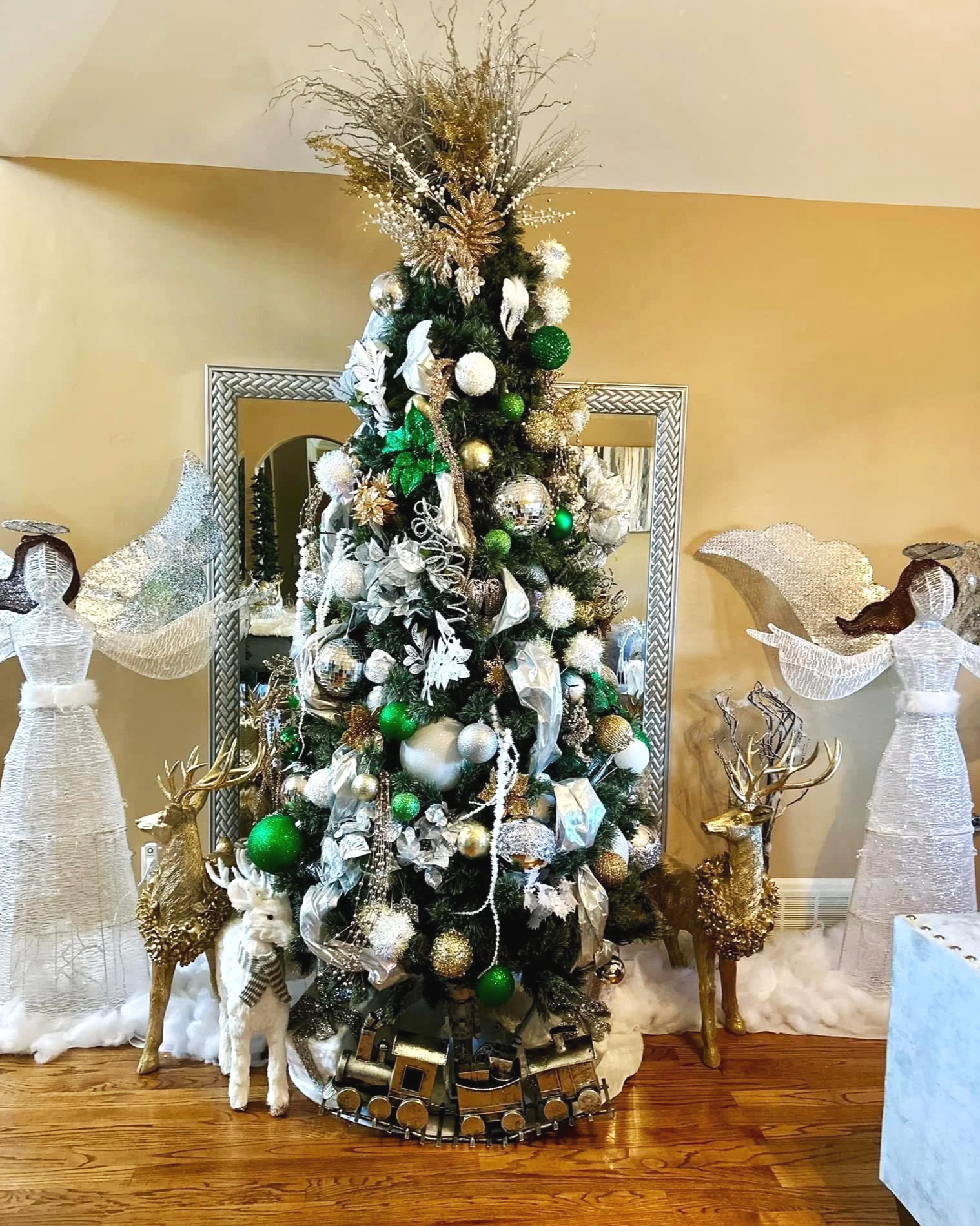 Decorated Christmas tree with silver, gold, and green ornaments, surrounded by Christmas angel and reindeer figures, with a mirror reflecting the scene.