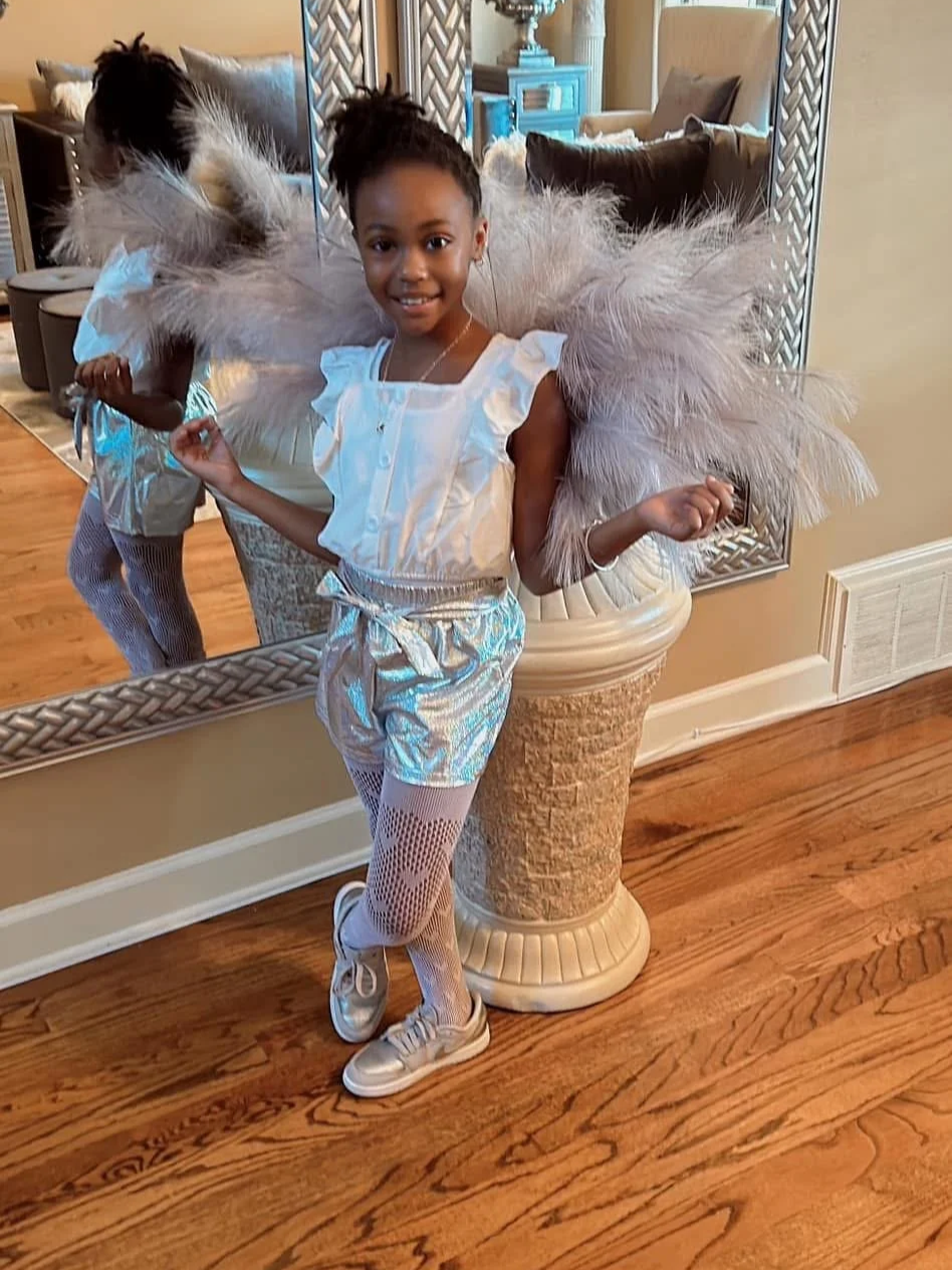 A young girl dressed in a shiny silver outfit with feathered wings, fishnet tights, and sneakers stands indoors next to a large mirror and tall decor with feathers.