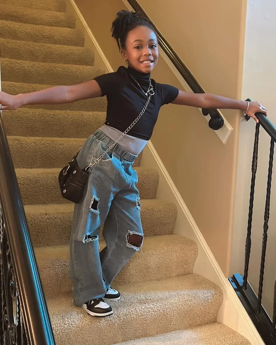 A young girl with styled hair, wearing a black t-shirt, distressed baggy jeans, and sneakers, posing on a carpeted staircase with her arms outstretched, smiling at the camera.