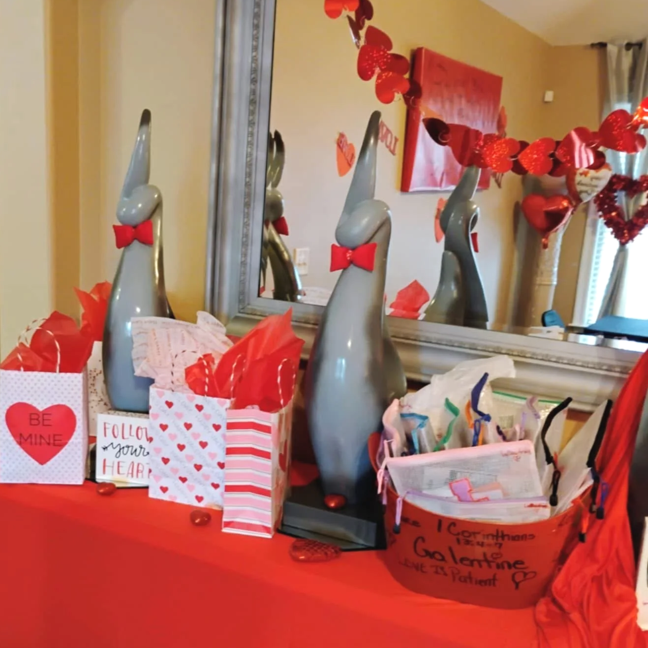 Valentine's Day decorations with red and white themed gift bags, cards, and paper napkins, red tissue paper, and bunny-shaped vases with red bow ties, all placed on a red tablecloth.