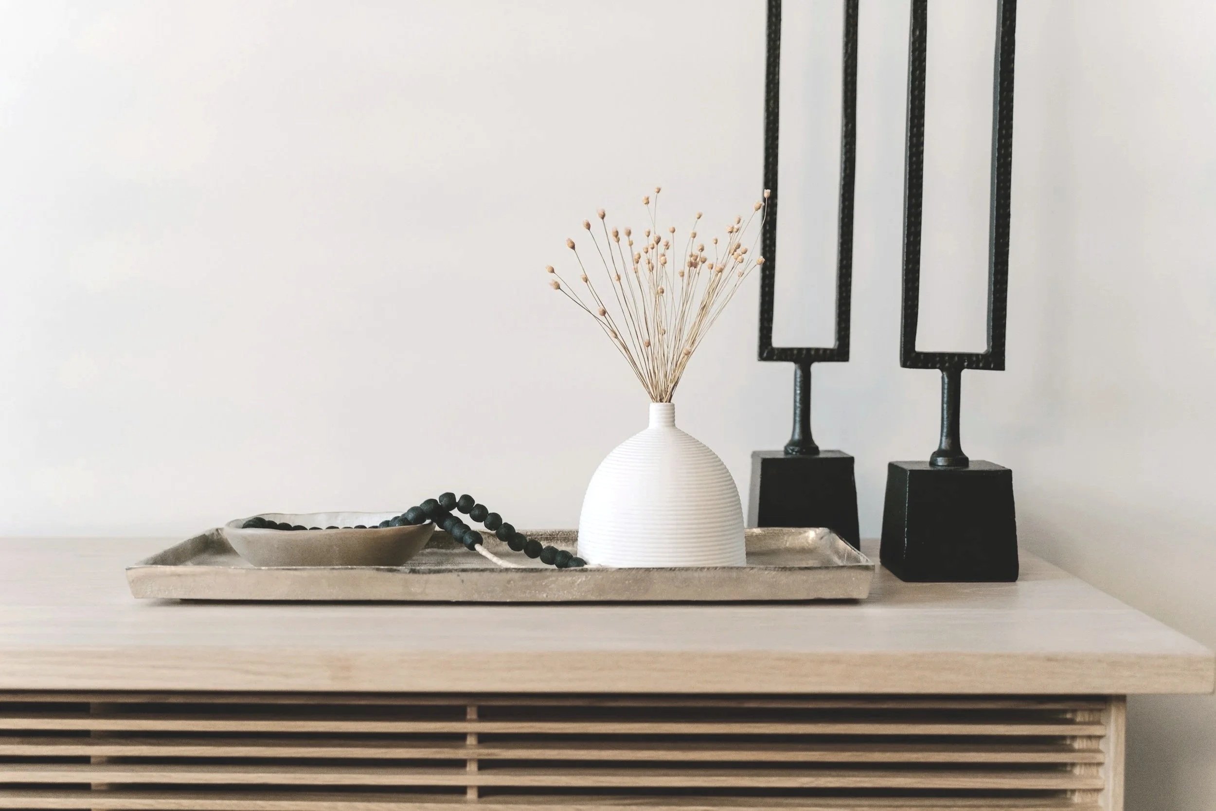 Minimalist decor on a wooden sideboard including a white ceramic vase with dried grass, a black beaded necklace on a beige tray, and black decorative frames against a white wall.