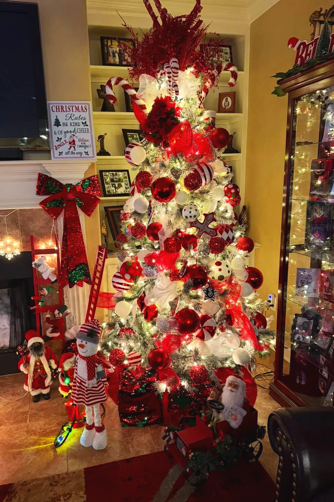 Decorated white Christmas tree with red and white ornaments, candy canes, ribbons, and lights in a cozy living room. Surrounding decorations include a snowman figure, presents, and holiday-themed ornaments.
