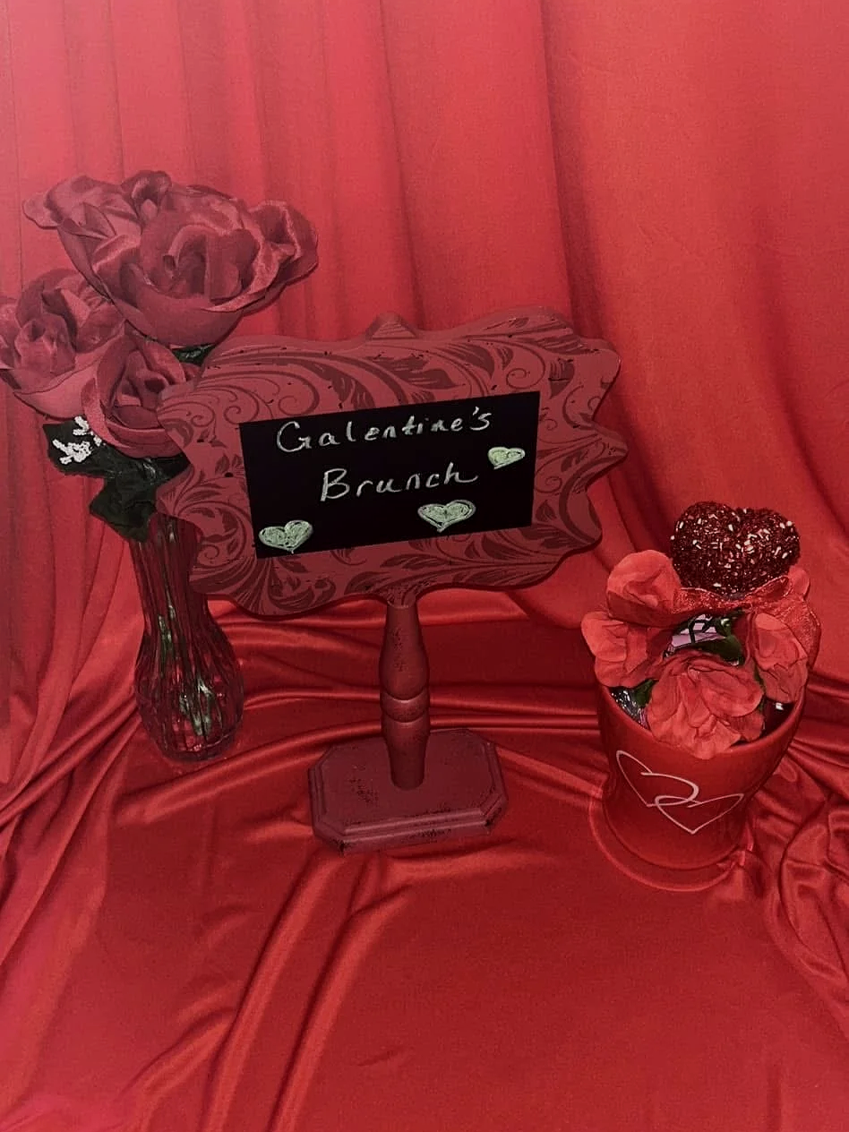 A Valentine's Day brunch display with a pink flower bouquet in a glass vase, a decorative sign reading 'Valentine's Brunch' with hearts, and a red potted plant with a glittery heart decoration, all arranged on red satin fabric.