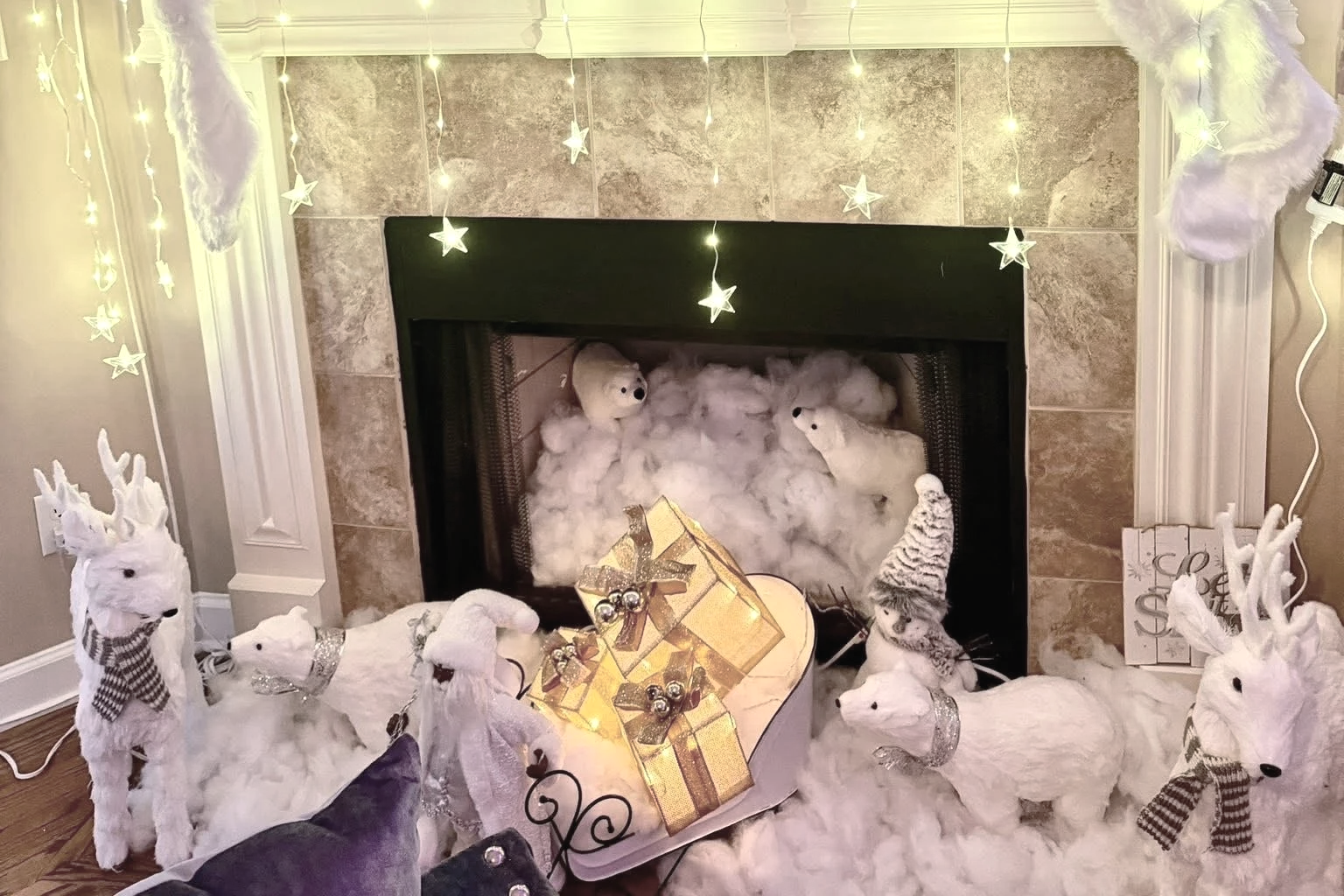 Festive holiday display with stuffed reindeer toys, a stack of wrapped gifts, string lights shaped like stars hanging above a fireplace, and cotton resembling snow, creating a cozy, wintery scene.