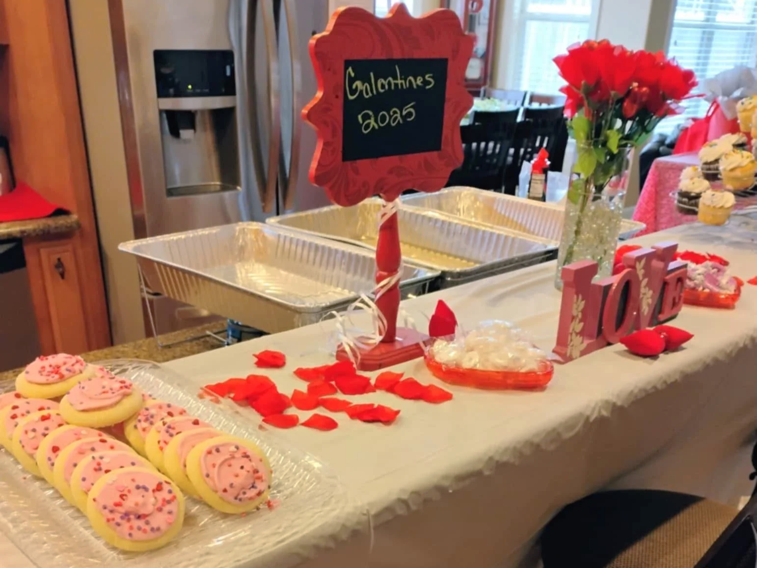 Valentine's Day dessert table with pink cookies, red rose petals, a bouquet of red flowers, love-themed decorations, and a sign indicating "Galentines 2023."