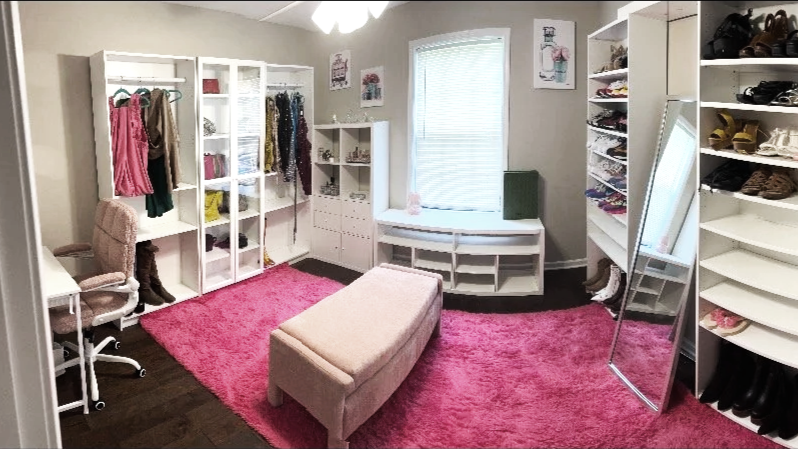 A walk-in closet with white shelving units, clothing, shoes, a pink area rug, a pink cushioned bench, a full-length mirror, and a window with curtains.