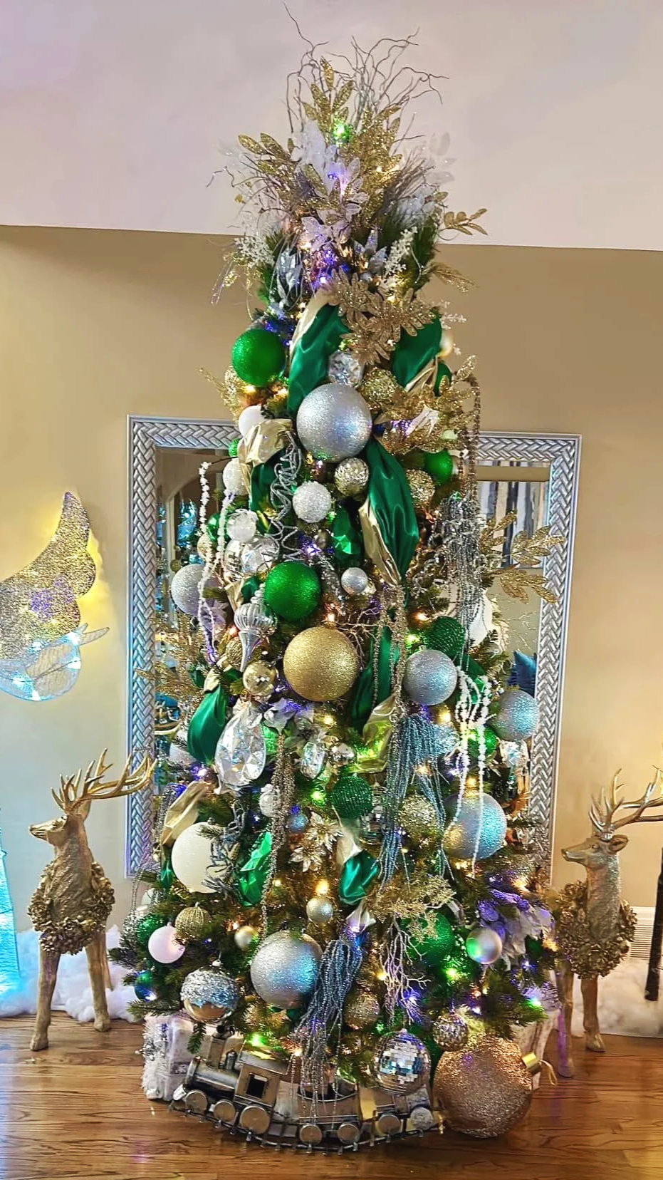 Decorated Christmas tree with gold, white, green ornaments, ribbons, and lights, flanked by two gold reindeer statues on a wooden floor.