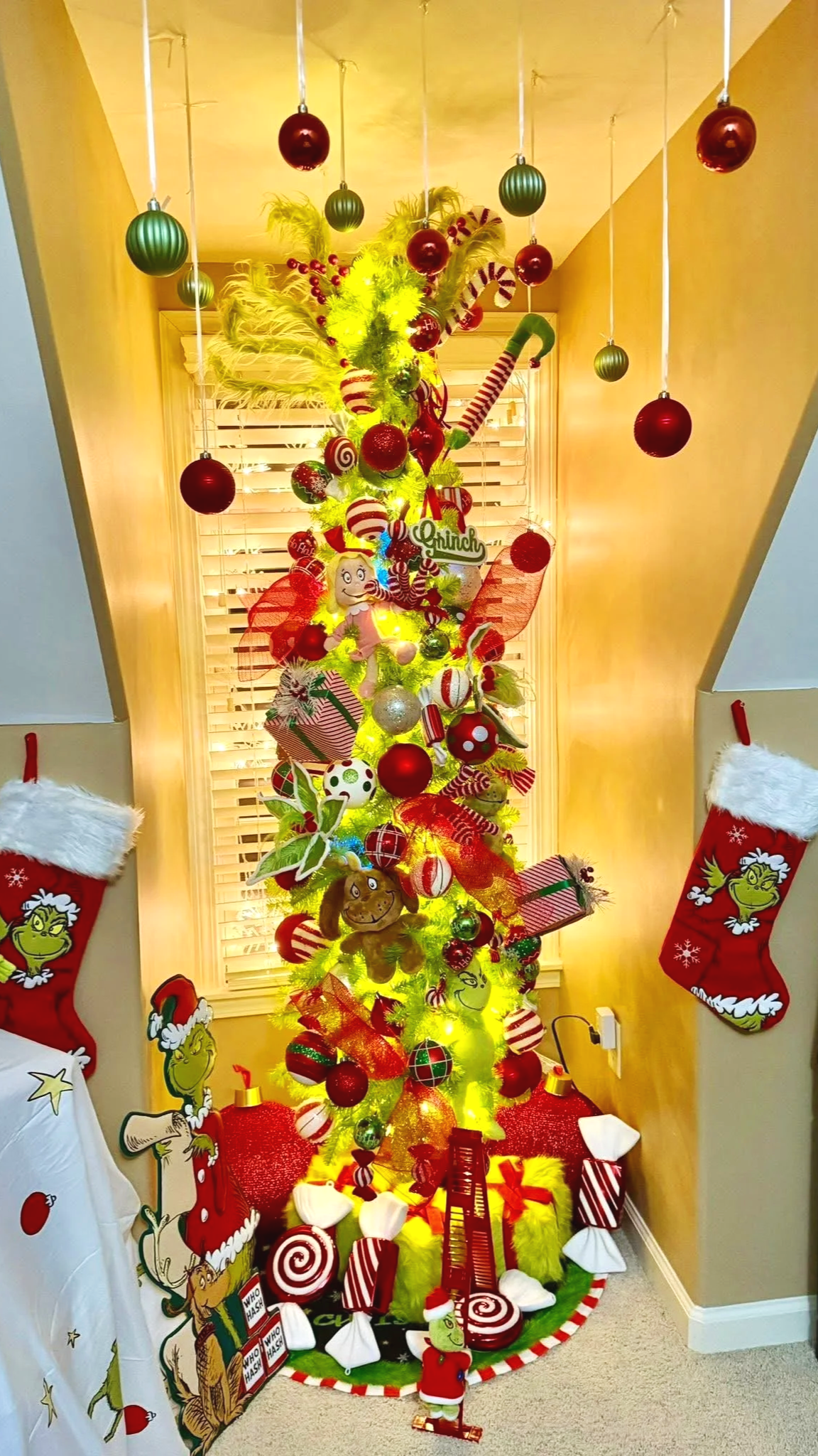 A brightly lit, decorated Christmas tree with red, green, and gold ornaments, candy cane decorations, plush toys, and lights. There are Christmas stockings on the wall with Grinch illustrations hanging above a bed with a Grinch-themed blanket.