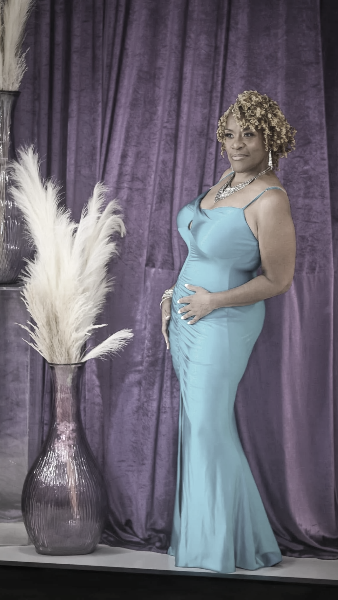 A woman in a light blue gown standing beside a large vase filled with pampas grass, with a purple curtain backdrop.