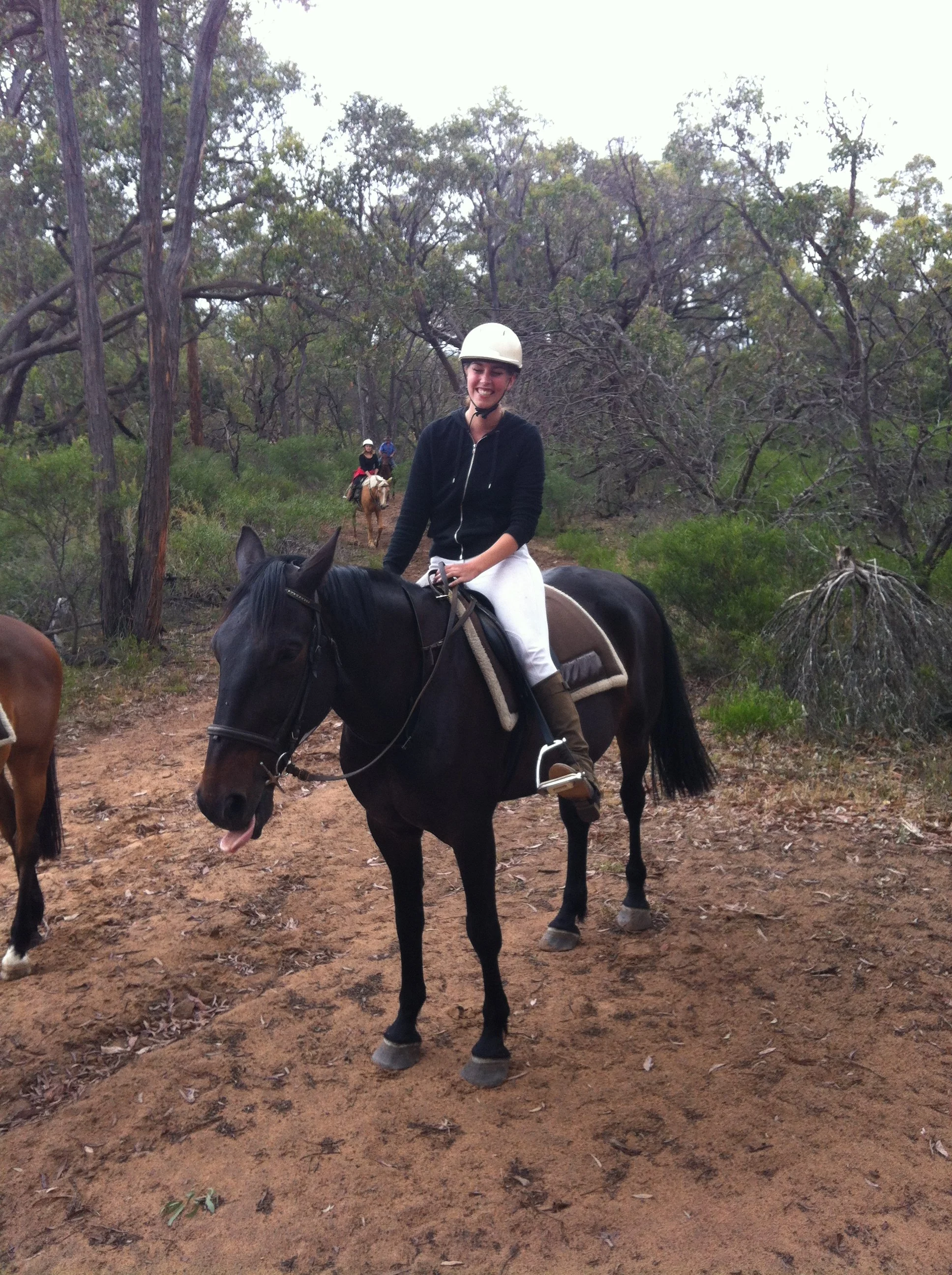 A woman smiling while riding a black horse on a dirt trail surrounded by trees, with two other riders visible in the background.