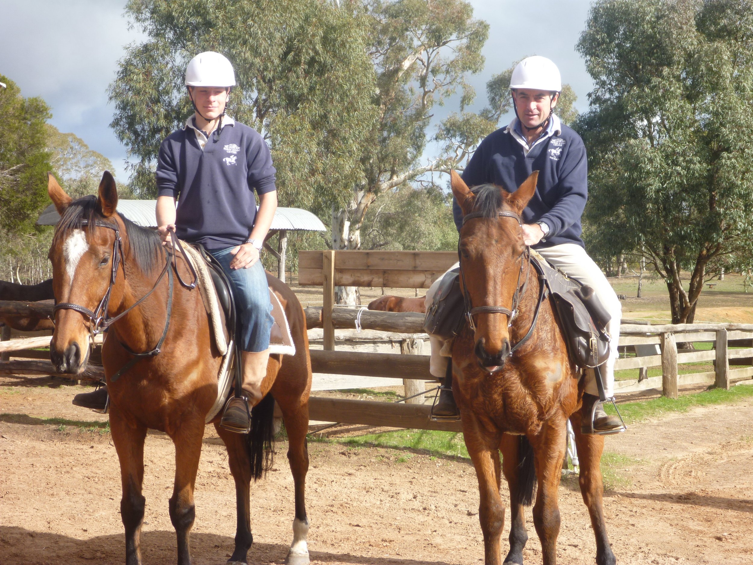 Two people riding horses outdoors on a sunny day, with trees and a wooden fence in the background.