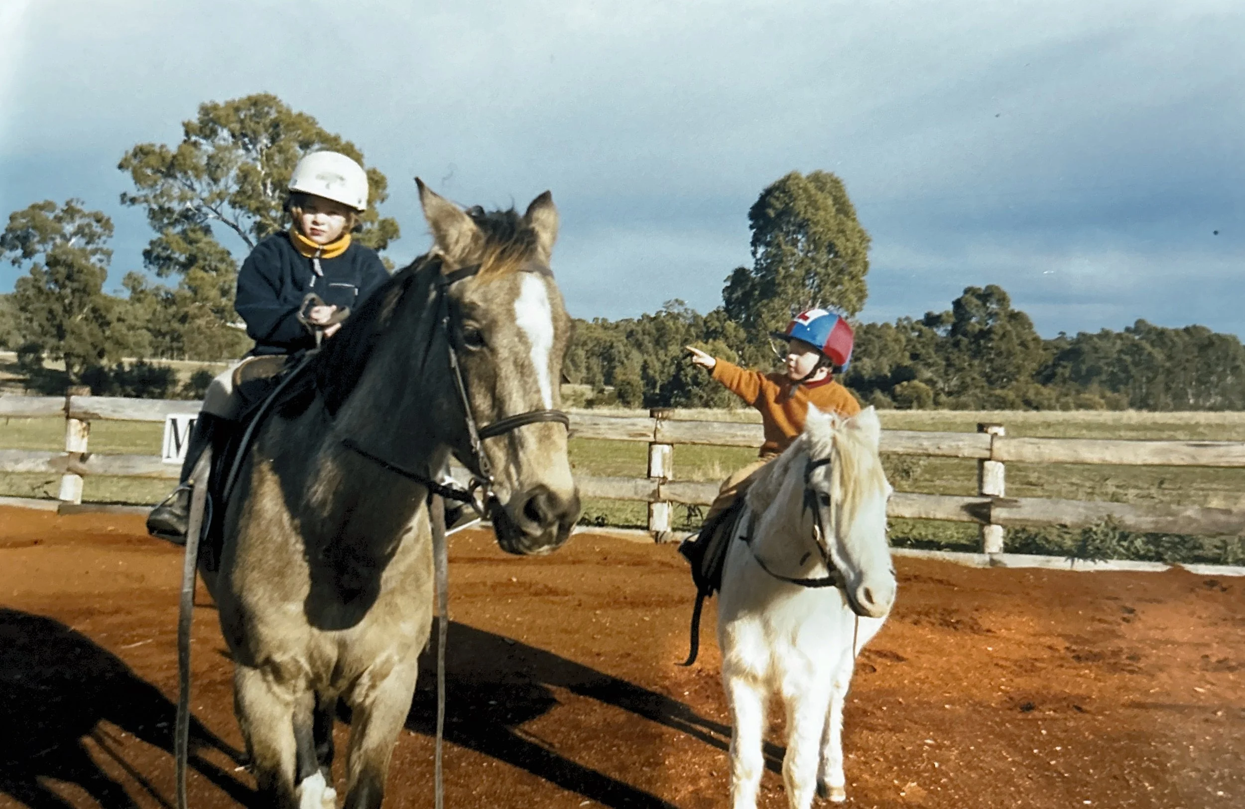 Two children riding horses on a dirt path near a wooden fence, with trees and a cloudy sky in the background. One child on the left is wearing a white helmet and navy jacket, and the other on the right is wearing a red and blue helmet and orange sweater.