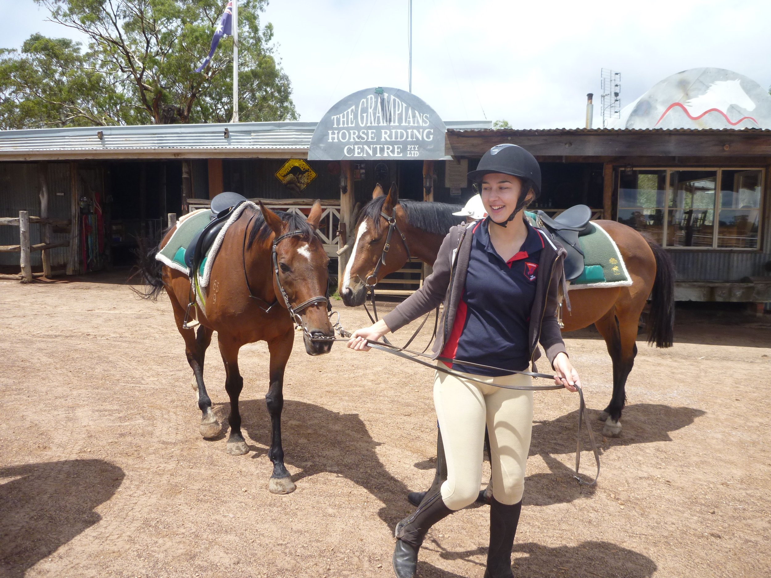 A woman in equestrian gear holding the reins of two saddled horses in front of a riding center sign, with a rustic building and trees in the background.