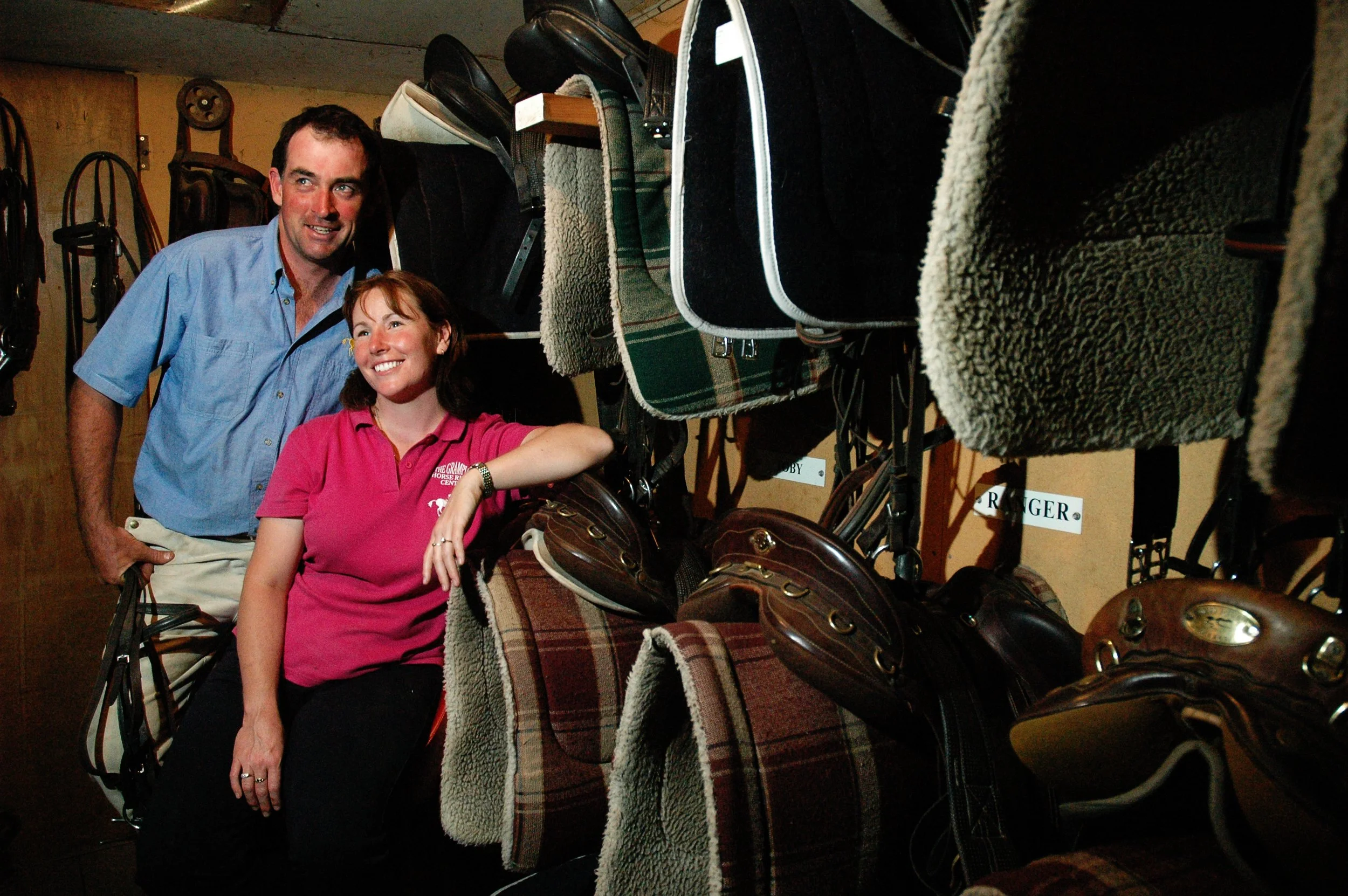 A man and a woman smiling inside a room filled with neatly arranged horse saddles and tack. Cameron and Sandra McDonald, directors of Grampians Horse Riding. 