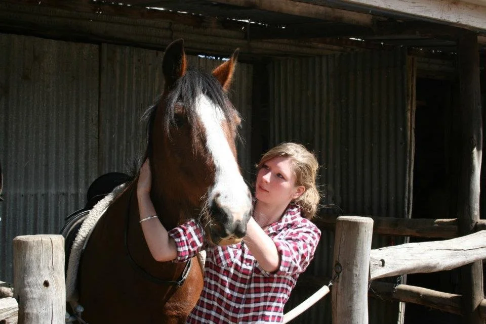 A young girl with blonde hair wearing a red and white checkered shirt gently touches the face of a brown and white horse inside a stable.
