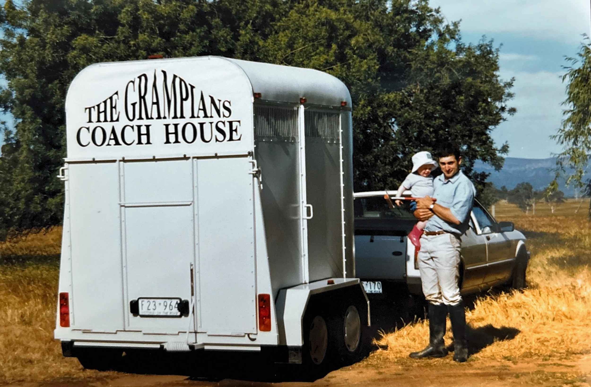 A man holding a child next to a white horse trailer with sign 'The Grampians Coach House' on it, in an outdoor rural area with trees and mountains in the background.