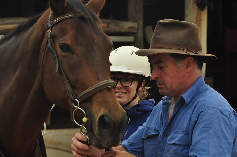 A woman in glasses and a man in a wide-brimmed hat are attending to a brown horse, with the woman smiling as she looks at the horse.