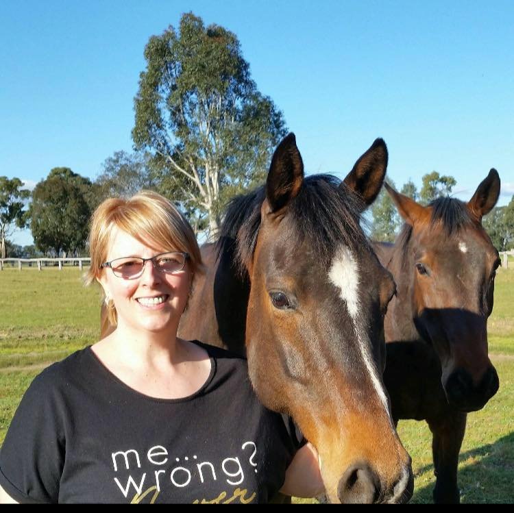 A woman with glasses and a black t-shirt smiling and standing outdoors next to two brown horses with white markings, on a green field with trees in the background.