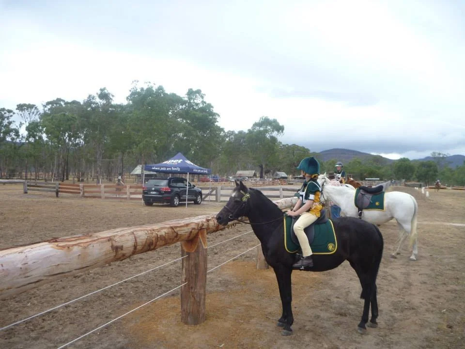 Children riding horses in an outdoor equestrian facility with trees, a car, and a tent in the background. Pony Club competition held at Grampians Horse Riding Centre.