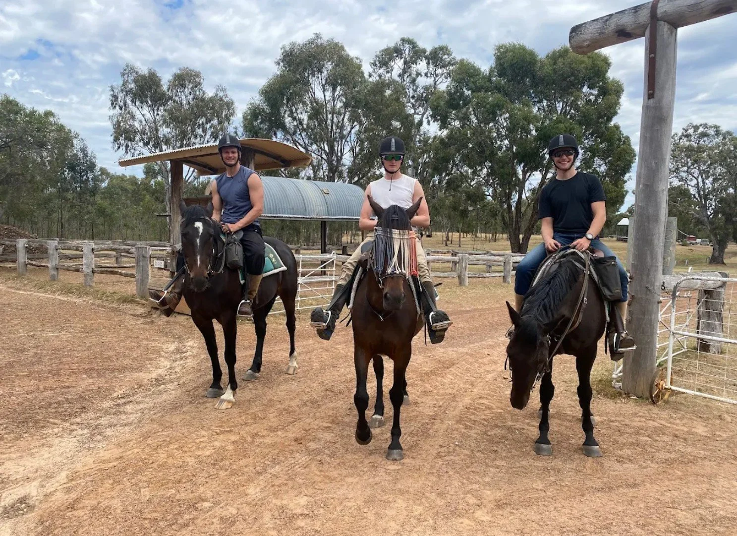 Three people riding horses on a dirt path at a farm or ranch, with trees and fencing in the background. All riders are wearing helmets.