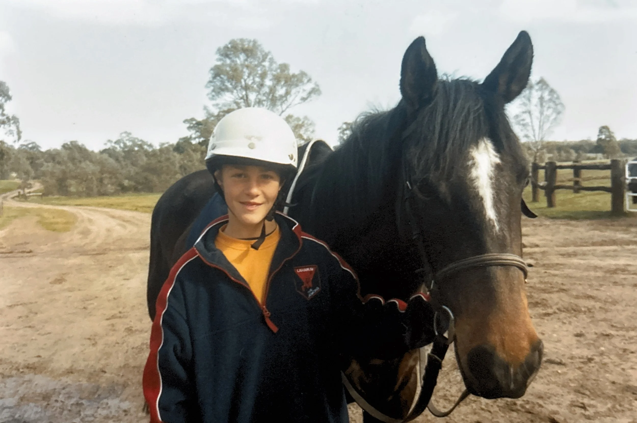 A young boy wearing a safety helmet and a dark jacket standing next to a brown and black horse, holding its bridle on a dirt path in a rural setting with trees and a wooden fence in the background.