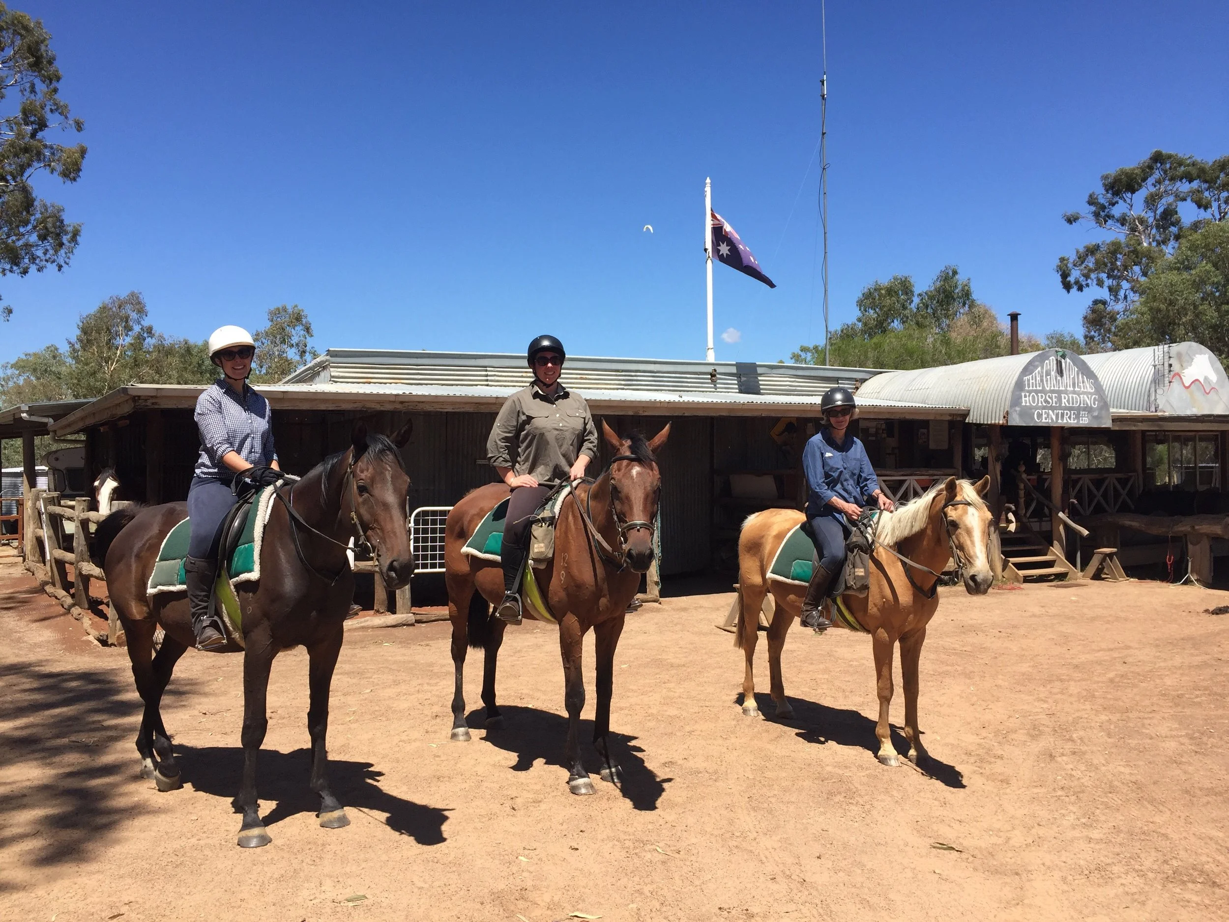 Three people riding horses in front of a rustic horse riding center under a blue sky.