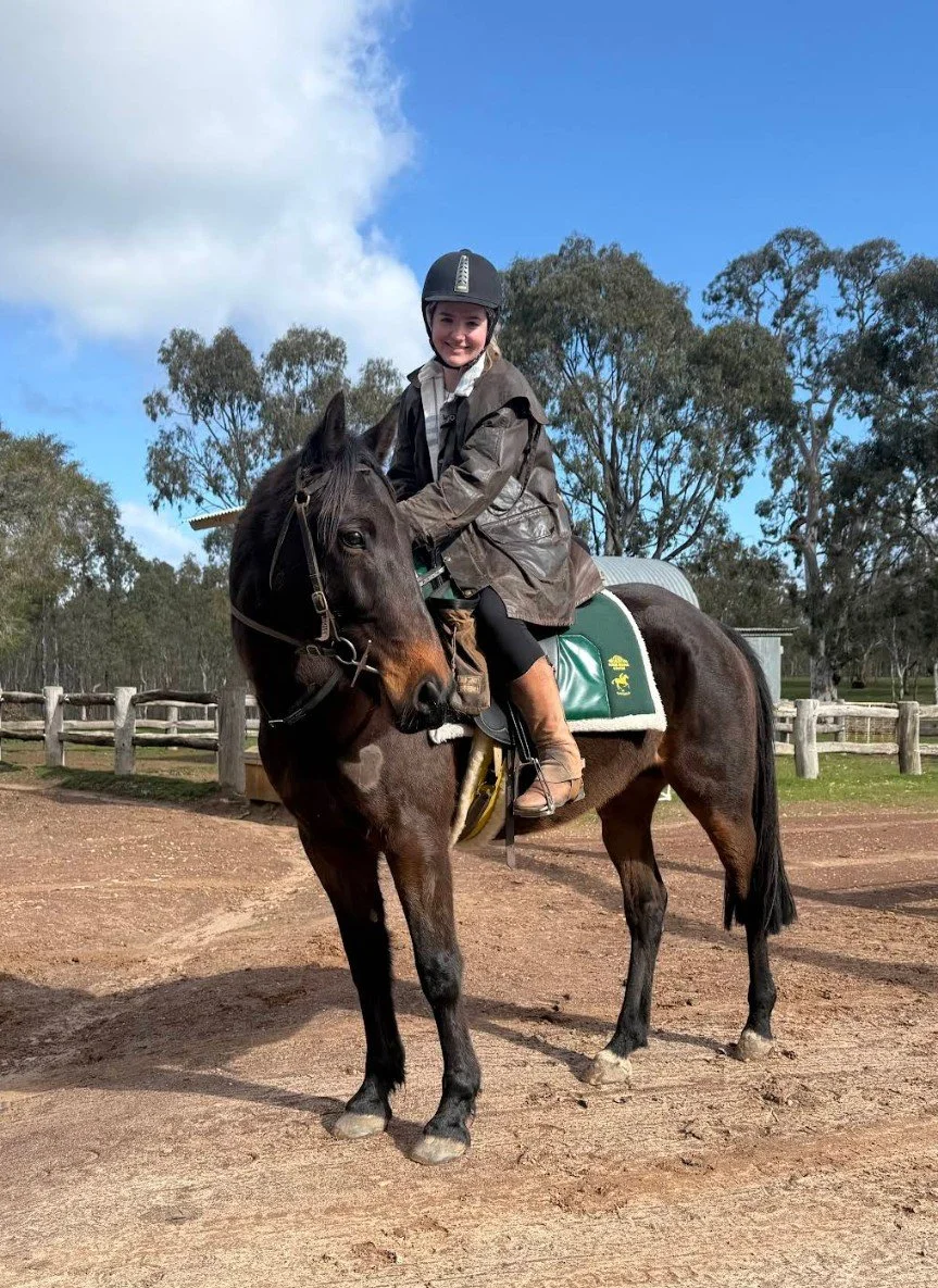 A young girl wearing a black riding helmet and brown riding jacket sitting on a brown horse with a green saddle pad outdoors on a dirt path, with trees and a blue partly cloudy sky in the background.
