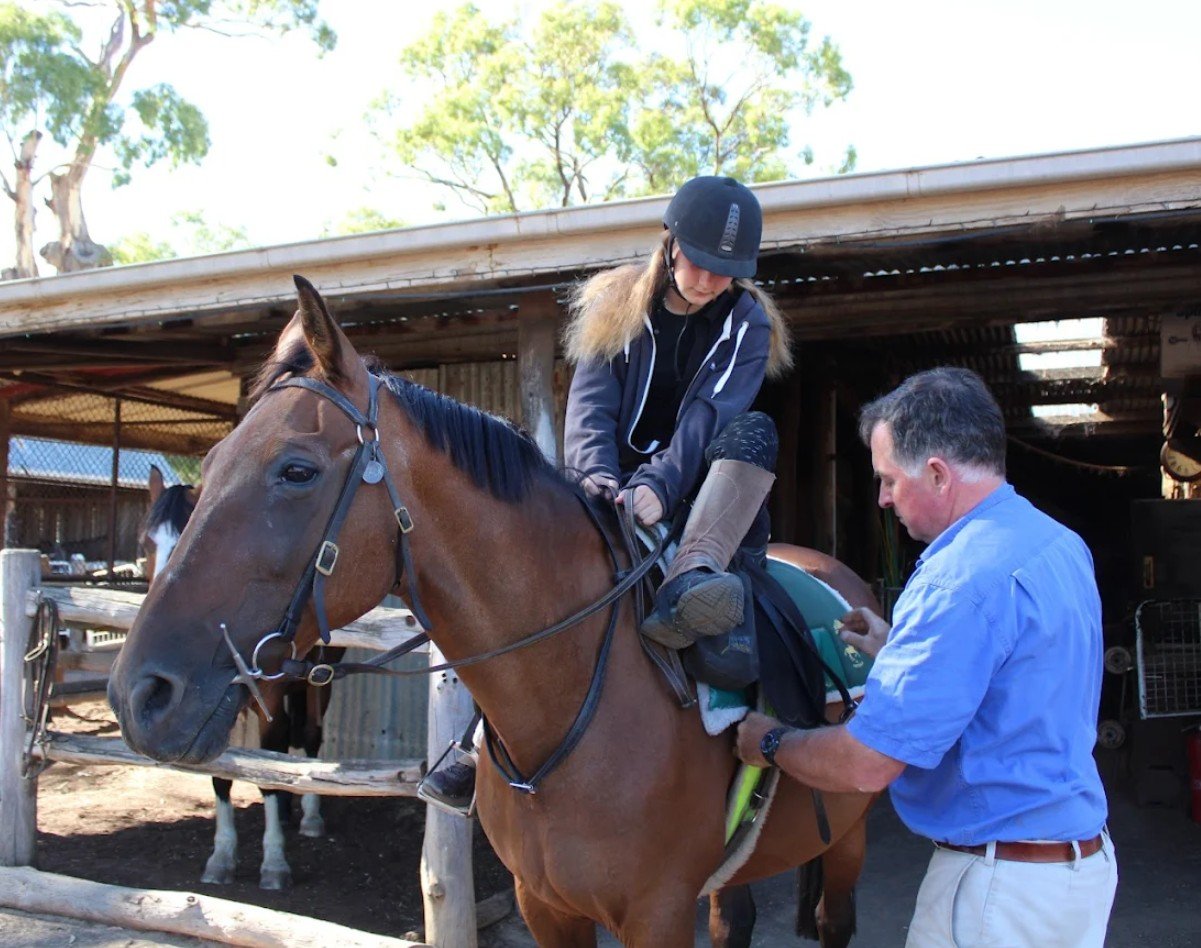 A young woman with long blonde hair, wearing a helmet, jacket, and boots, sits on a brown horse while a man in a blue shirt helps her get ready. They are outside near a wooden stable, with trees in the background.
