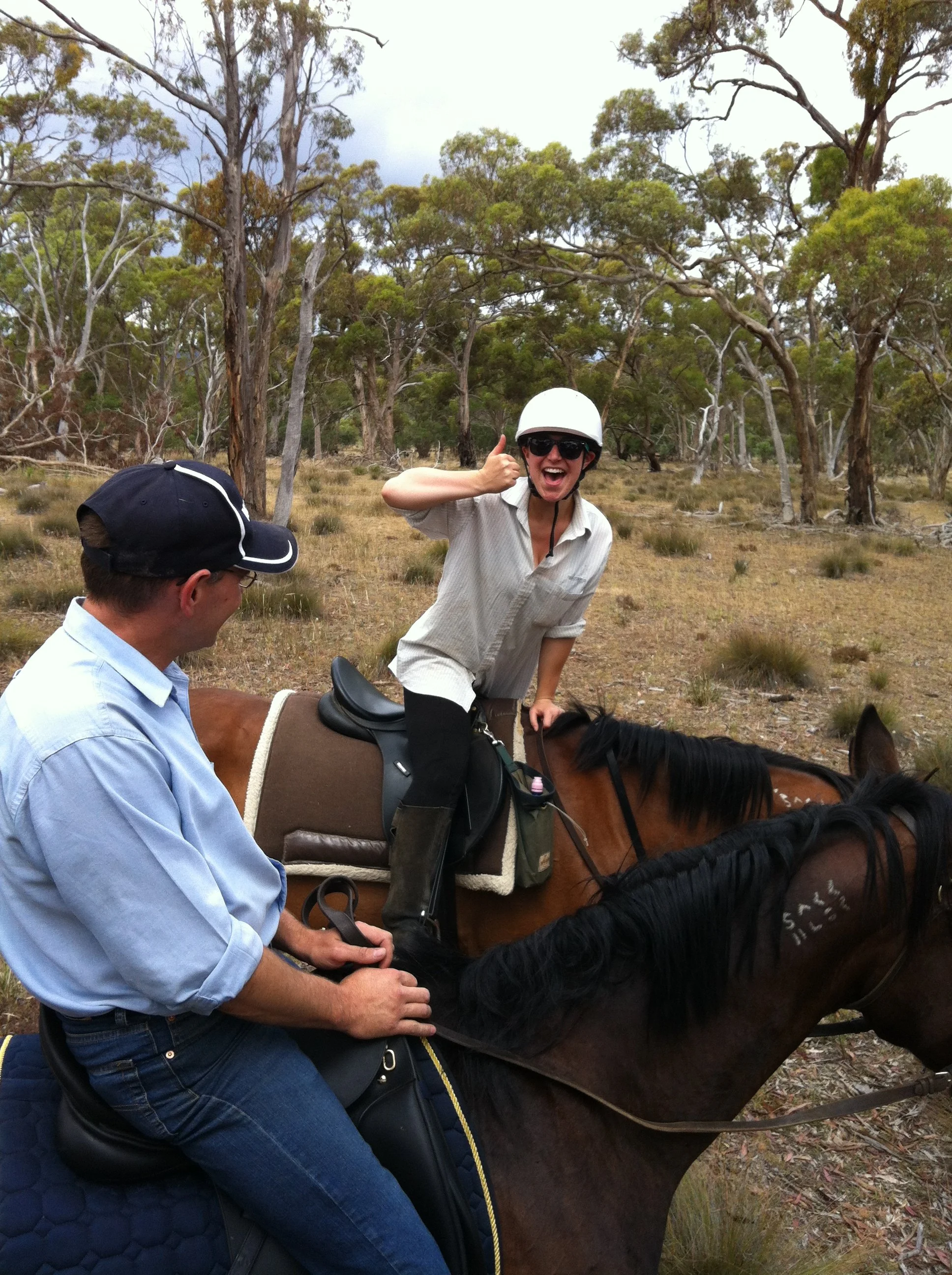 A woman riding a horse in a wooded outdoor area, smiling and giving a thumbs-up, with a man guiding the horse.