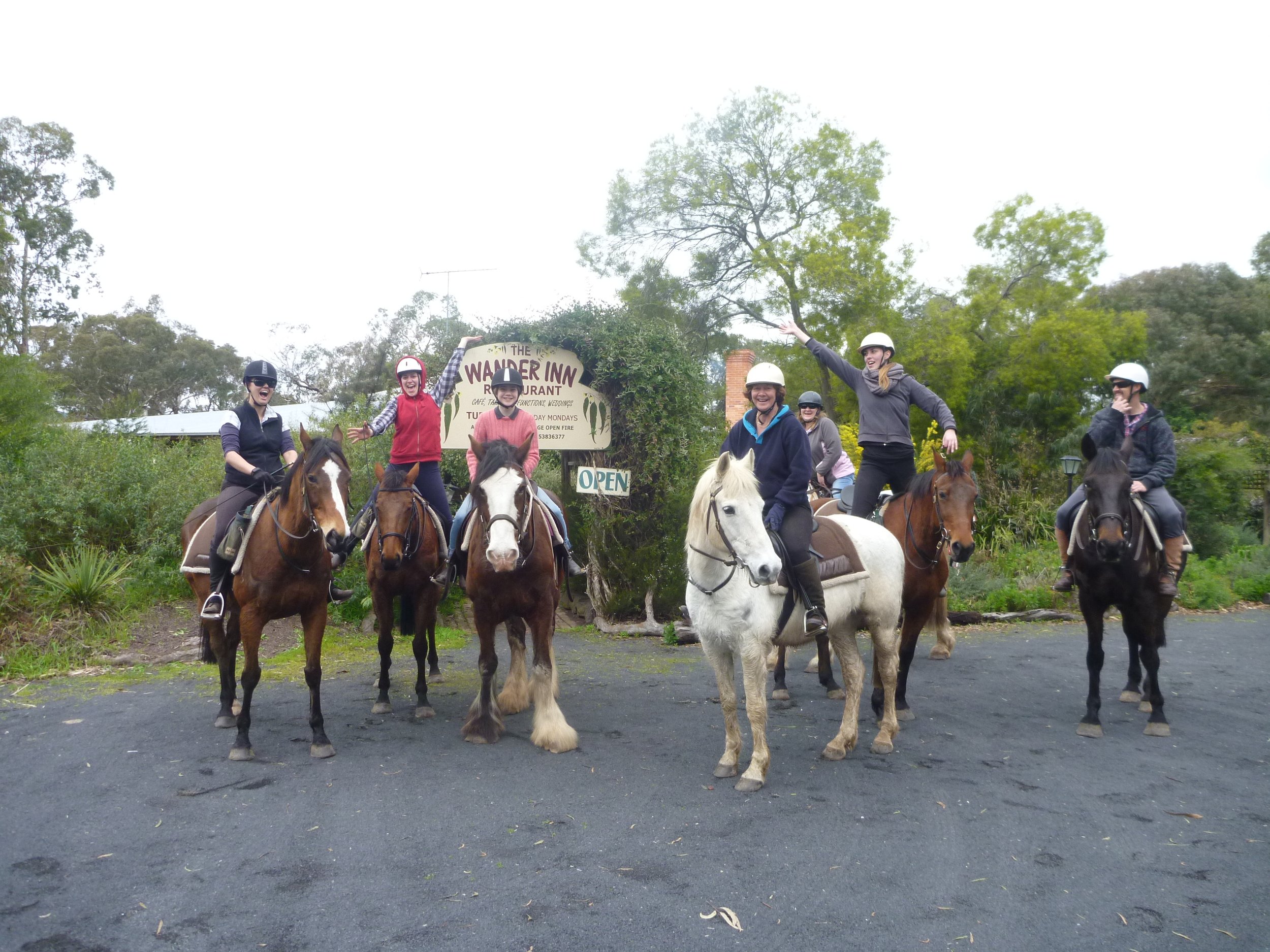 Six women wearing riding helmets riding horses and smiling, with some waving or making gestures, outdoors near greenery and trees. Local pub ride to The Wander Inn, Wartook Valley, Victoria.