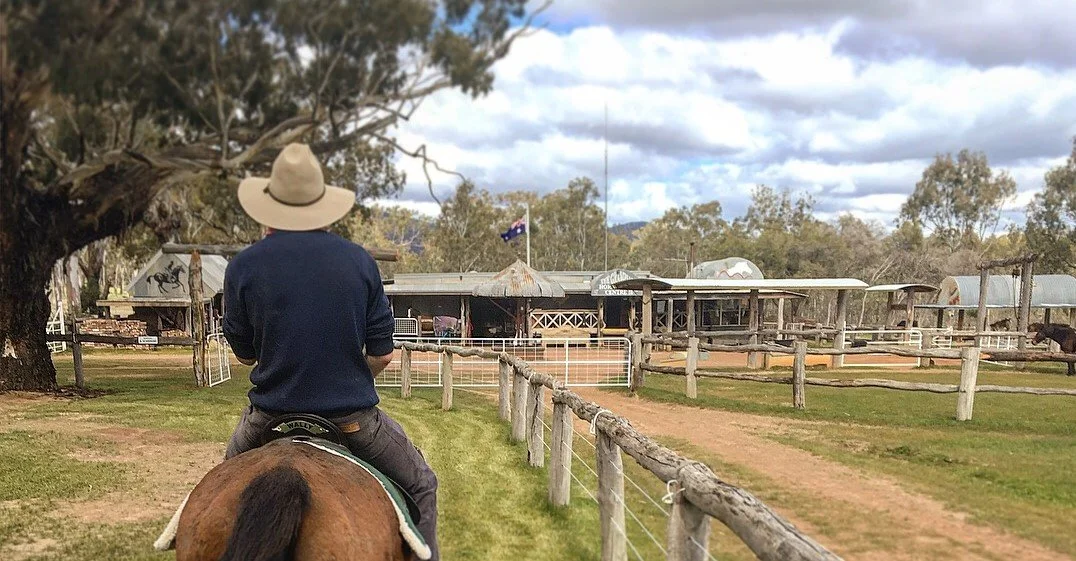 A person wearing a hat riding a horse on a farm, with a rustic barn and trees in the background under a cloudy sky.