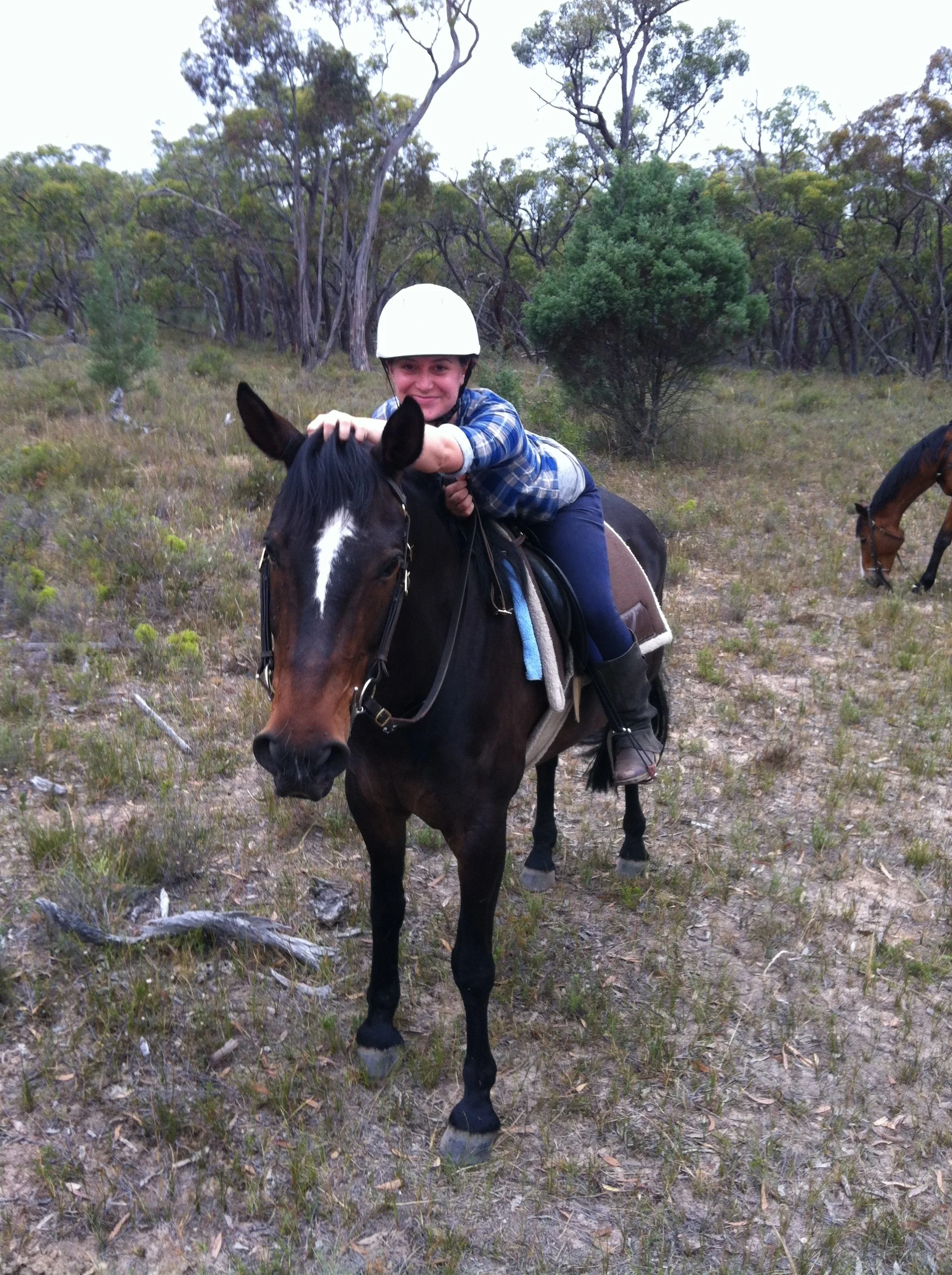 Person riding a brown and black horse in a natural outdoor setting with trees and grass, smiling and wearing a white helmet.