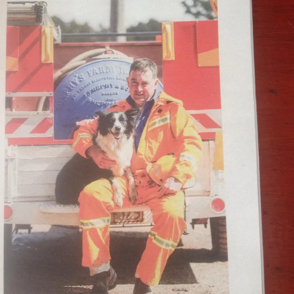 A firefighter in an orange uniform holding a black and white dog, sitting on the back of a fire truck. Weekly Times article on Cameron McDonald, fire plan for horse owners. 