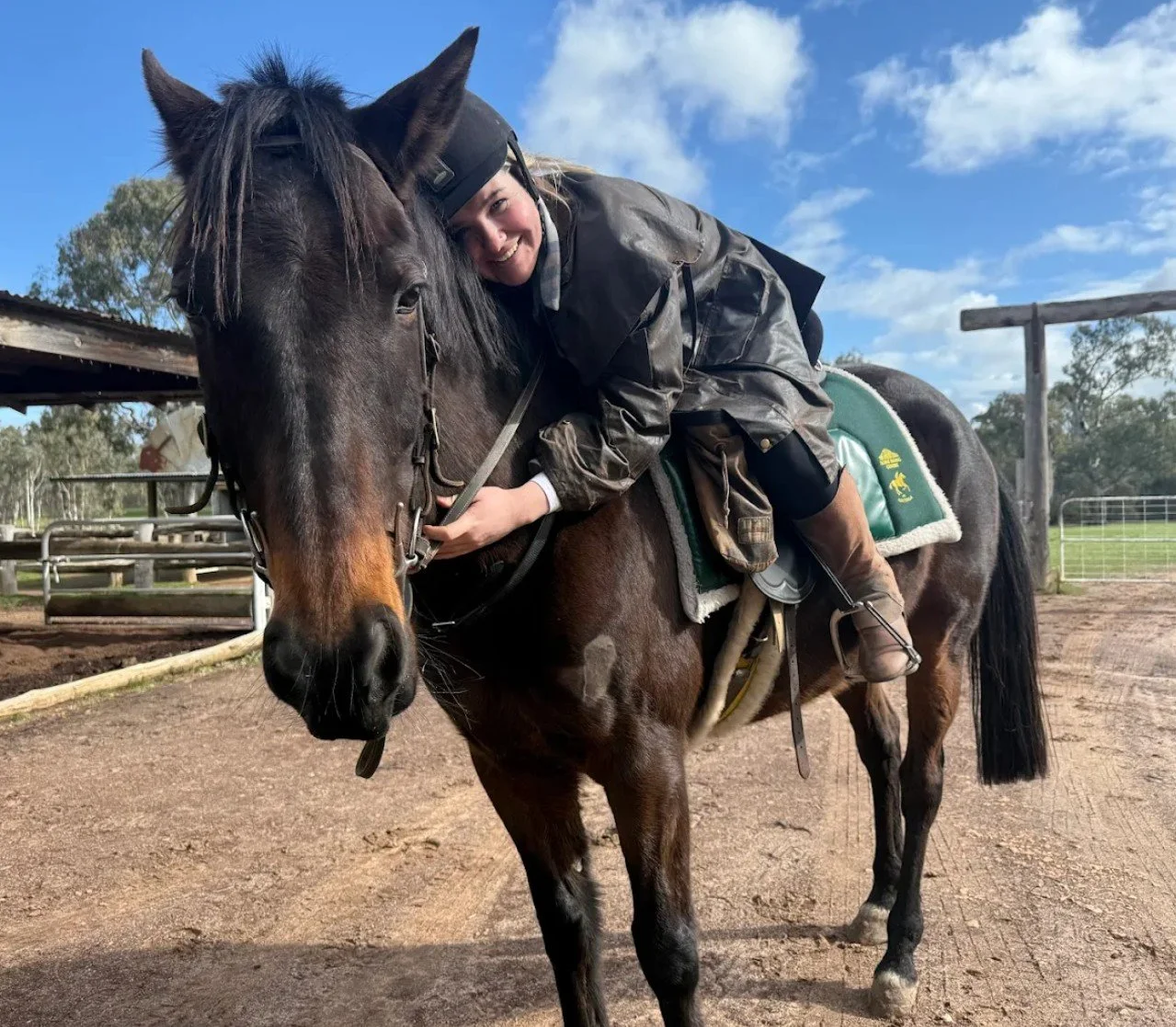 A woman in riding gear hugging a dark brown horse in an outdoor stable area with a blue sky and clouds in the background.