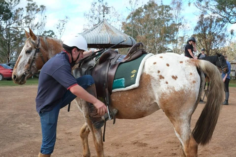 A man in a helmet adjusting the strap on a saddle on a brown and white spotted horse at a riding area with trees and other riders in the background.
