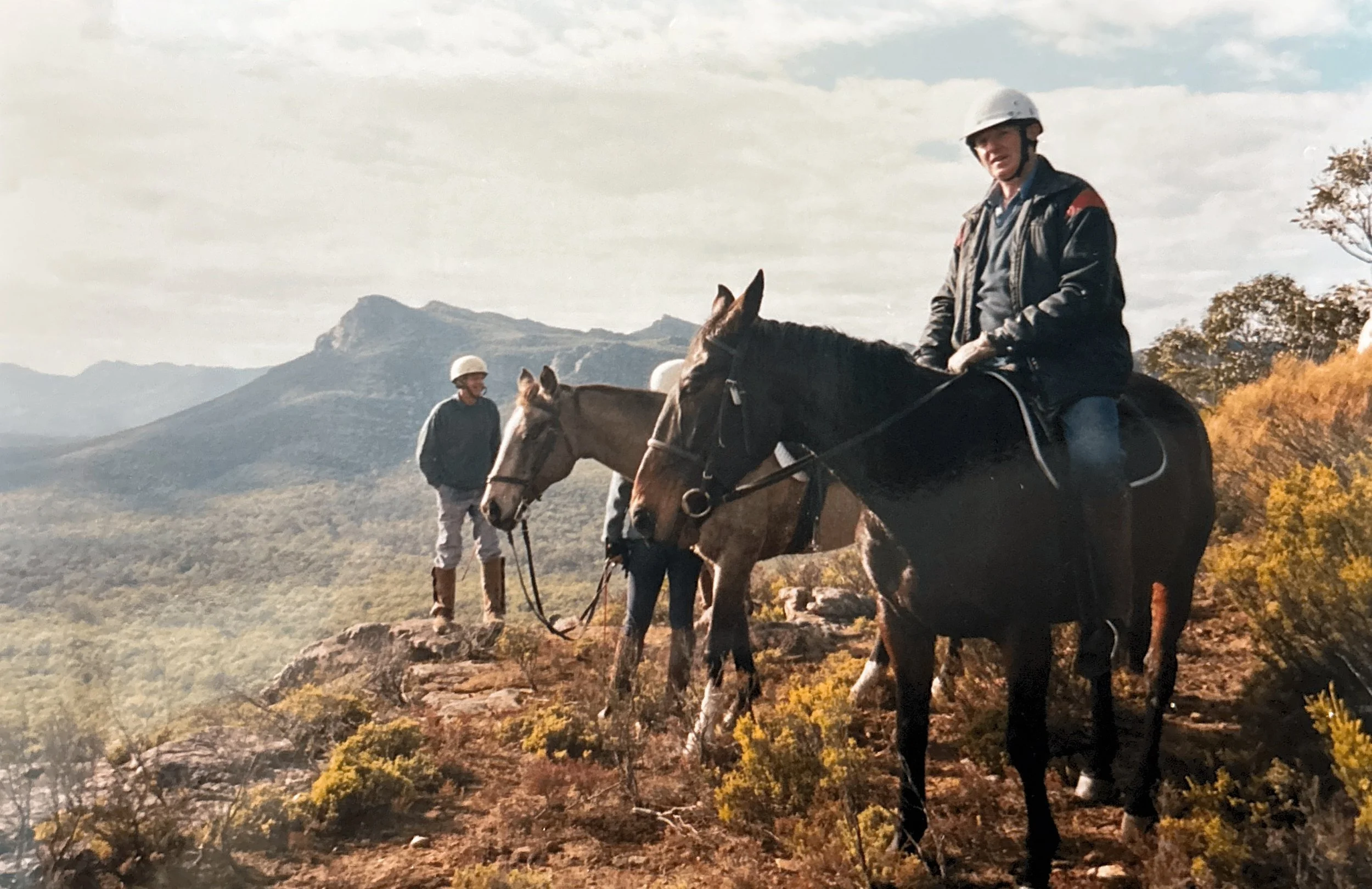 Two men on horseback atop a rocky hill with a mountain and cloudy sky in the background, surrounded by shrubs and trees with autumn foliage.