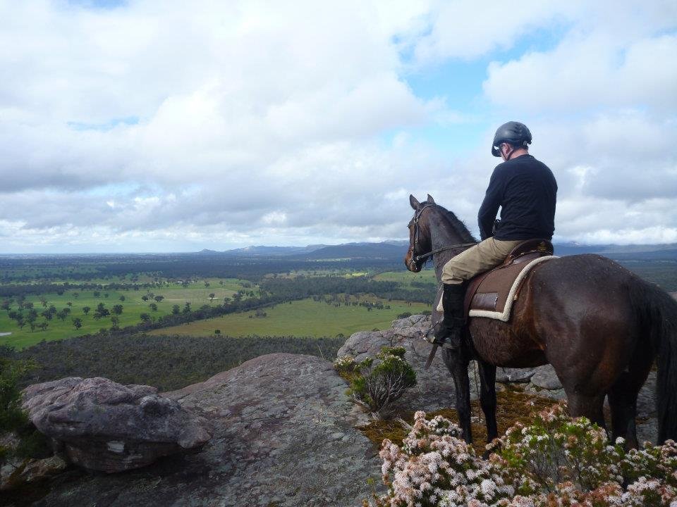 A person riding a horse on a rocky hilltop overlooking a green valley with trees and open fields, under a cloudy sky.