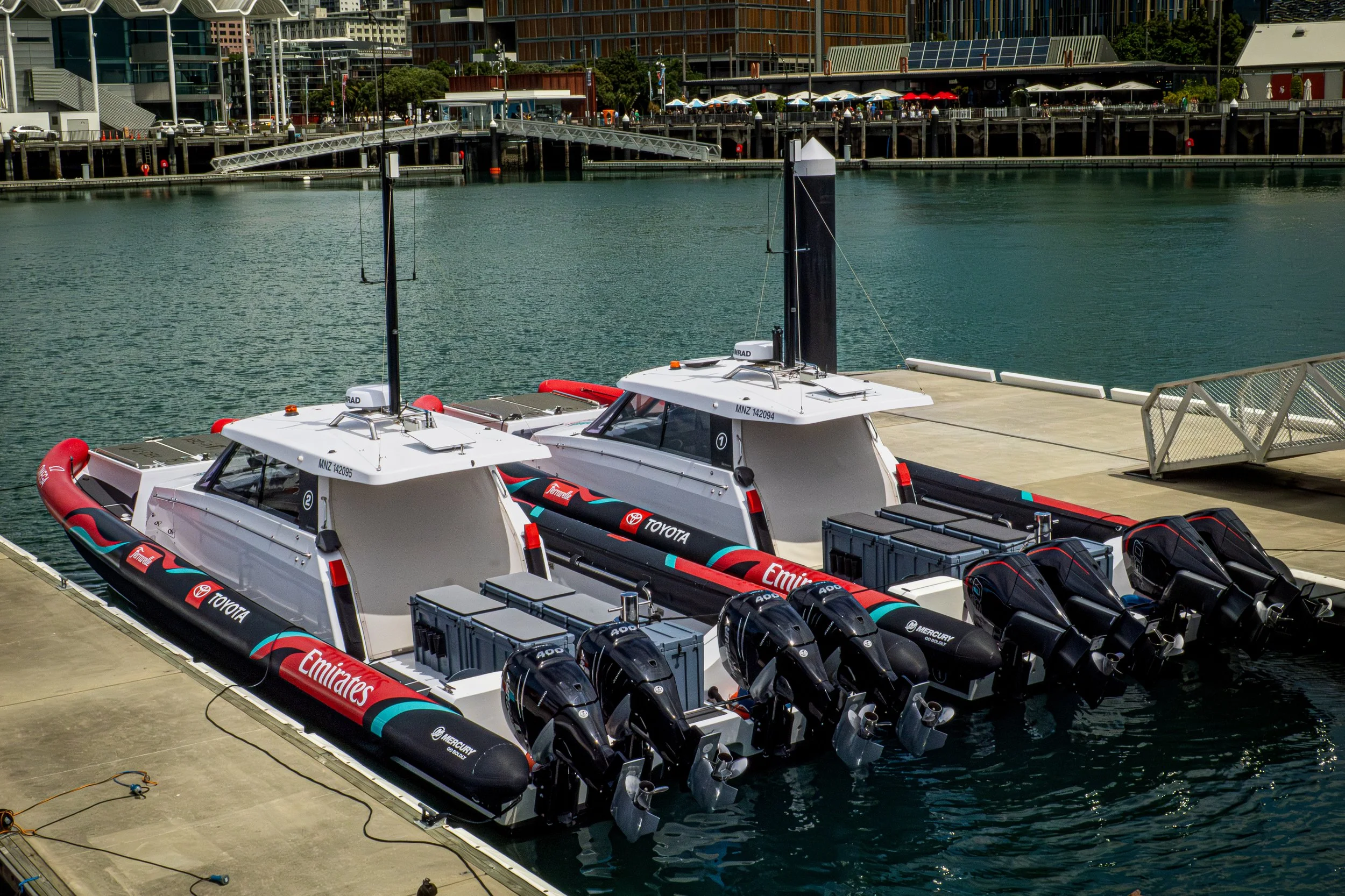 Two high-performance race boats with multiple engines docked at a marina, with a cityscape and waterfront in the background.