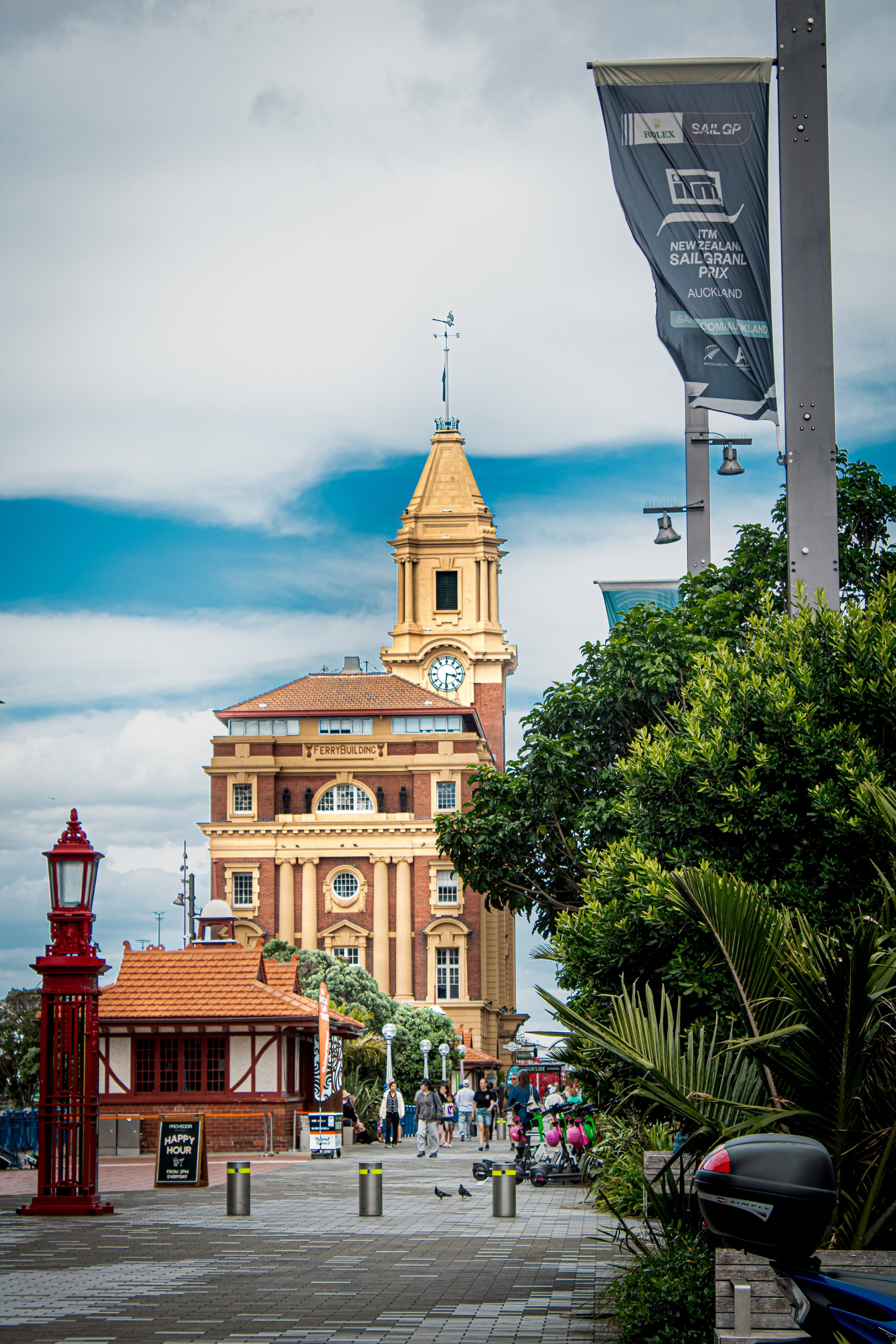 A historic building with a clock tower, labeled 'FERRY BUILDING,' is seen in an urban area with people walking, greenery, streetlights, and banners, under a cloudy sky.