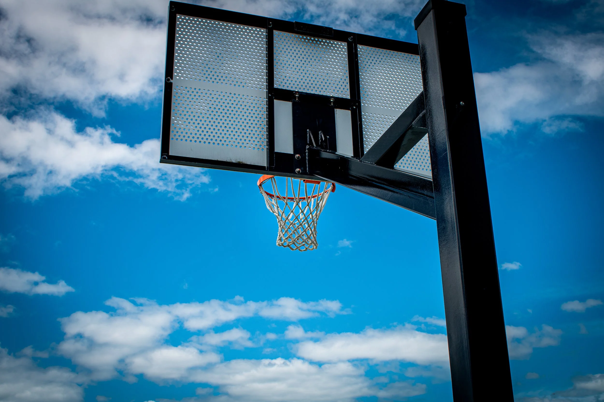 A basketball hoop with a black pole and backboard against a partly cloudy sky.