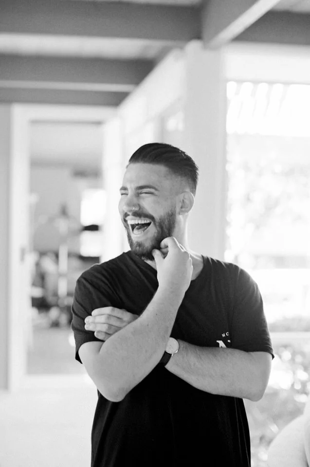 A young man with a beard and short hair, smiling and laughing, standing indoors near a window, with arms crossed and hand touching his chin.