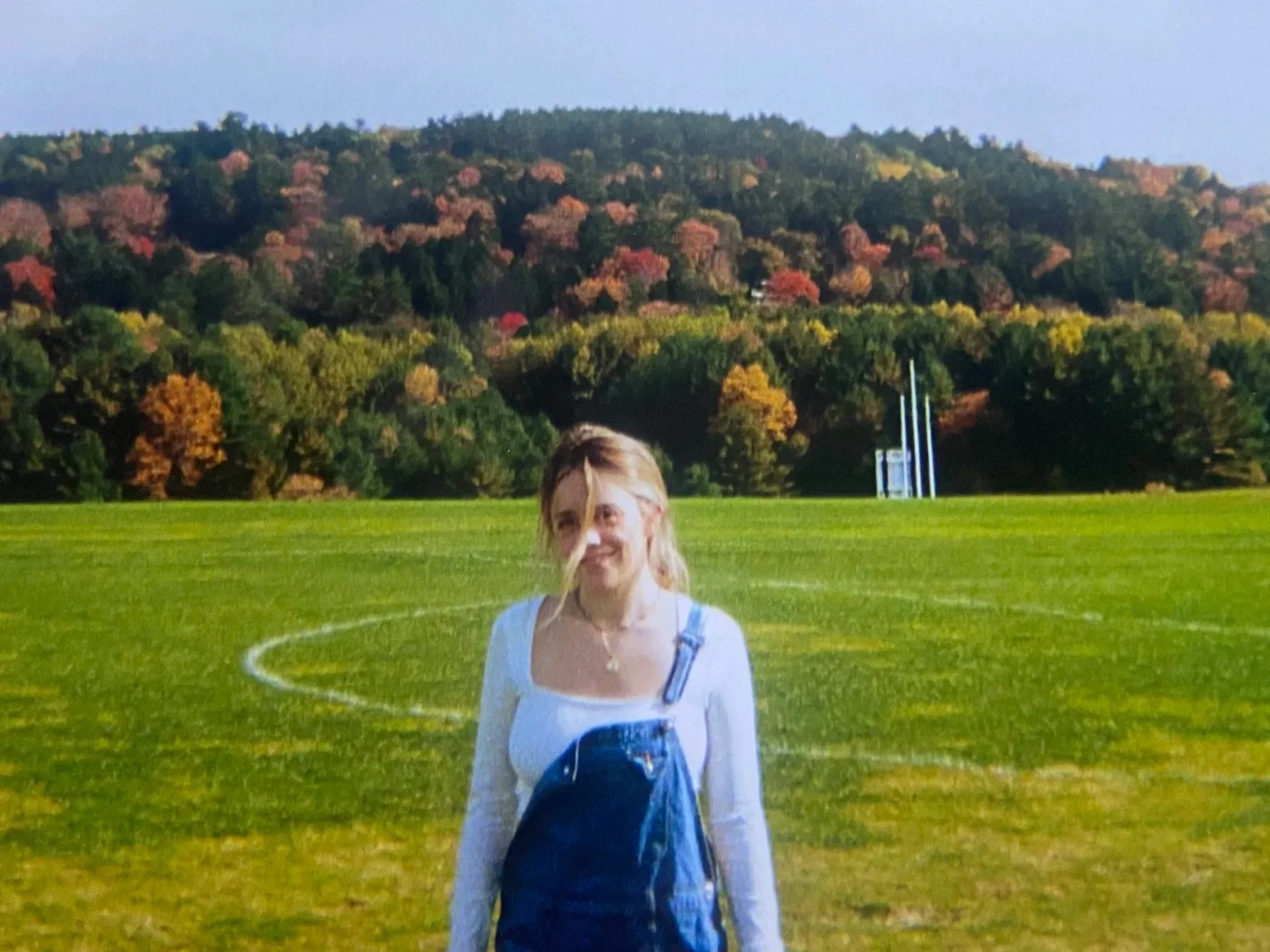 A smiling young woman with blonde hair stands on a green football field with a hill covered in trees with fall foliage in the background.